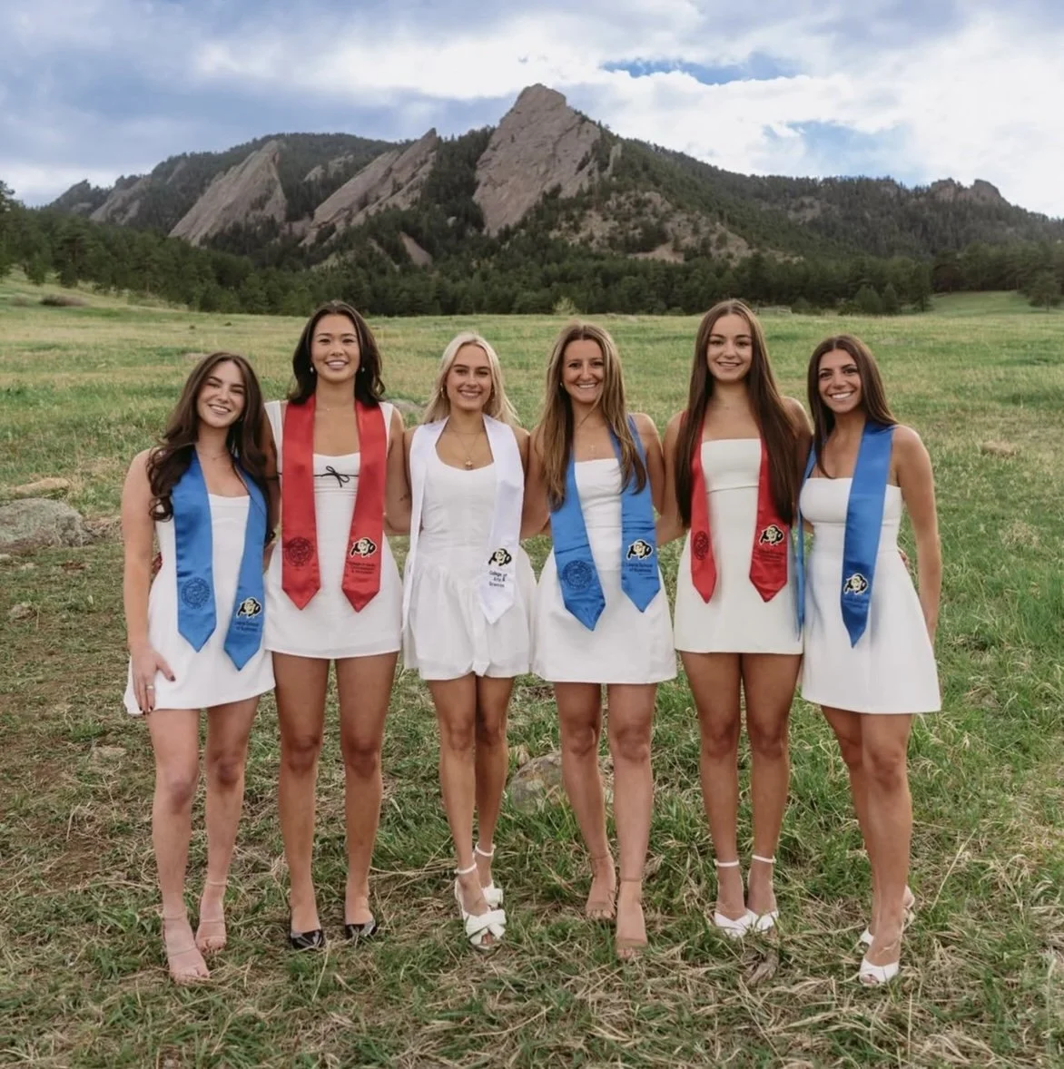 Six young women standing outdoors in a grassy field with mountains in the background, wearing white dresses and graduation sashes in red, blue, and white.