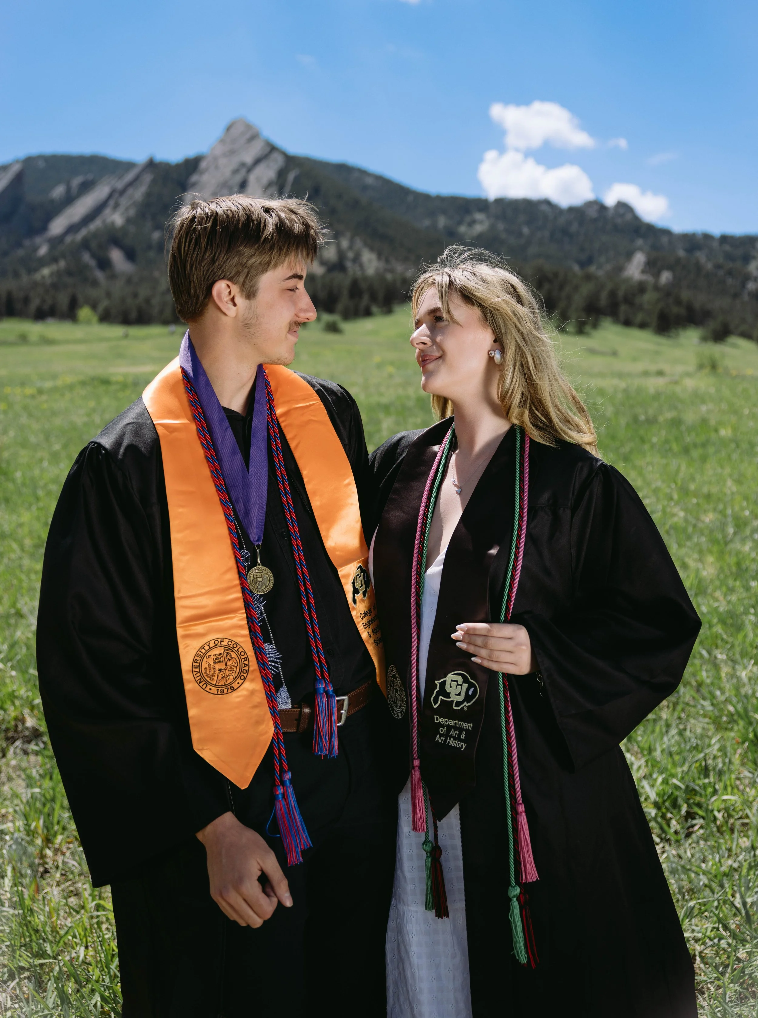CU Boulder couple graduates in caps and gowns posing together in green field