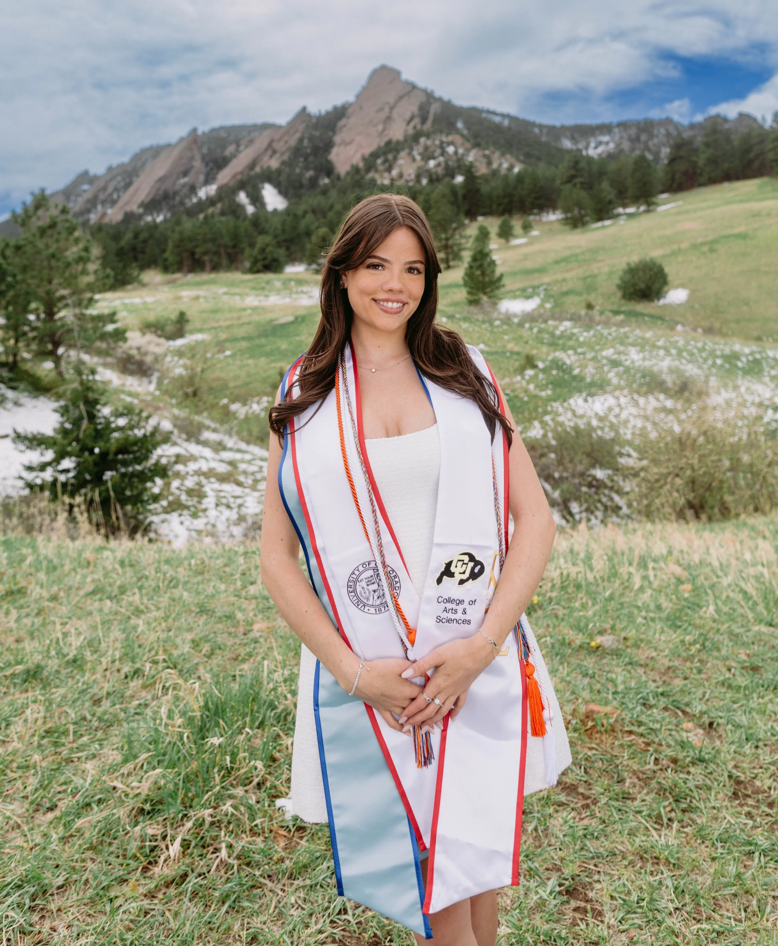 A young woman standing outdoors in a grassy field with mountains in the background, wearing a graduation stole from the University of Colorado Boulder.