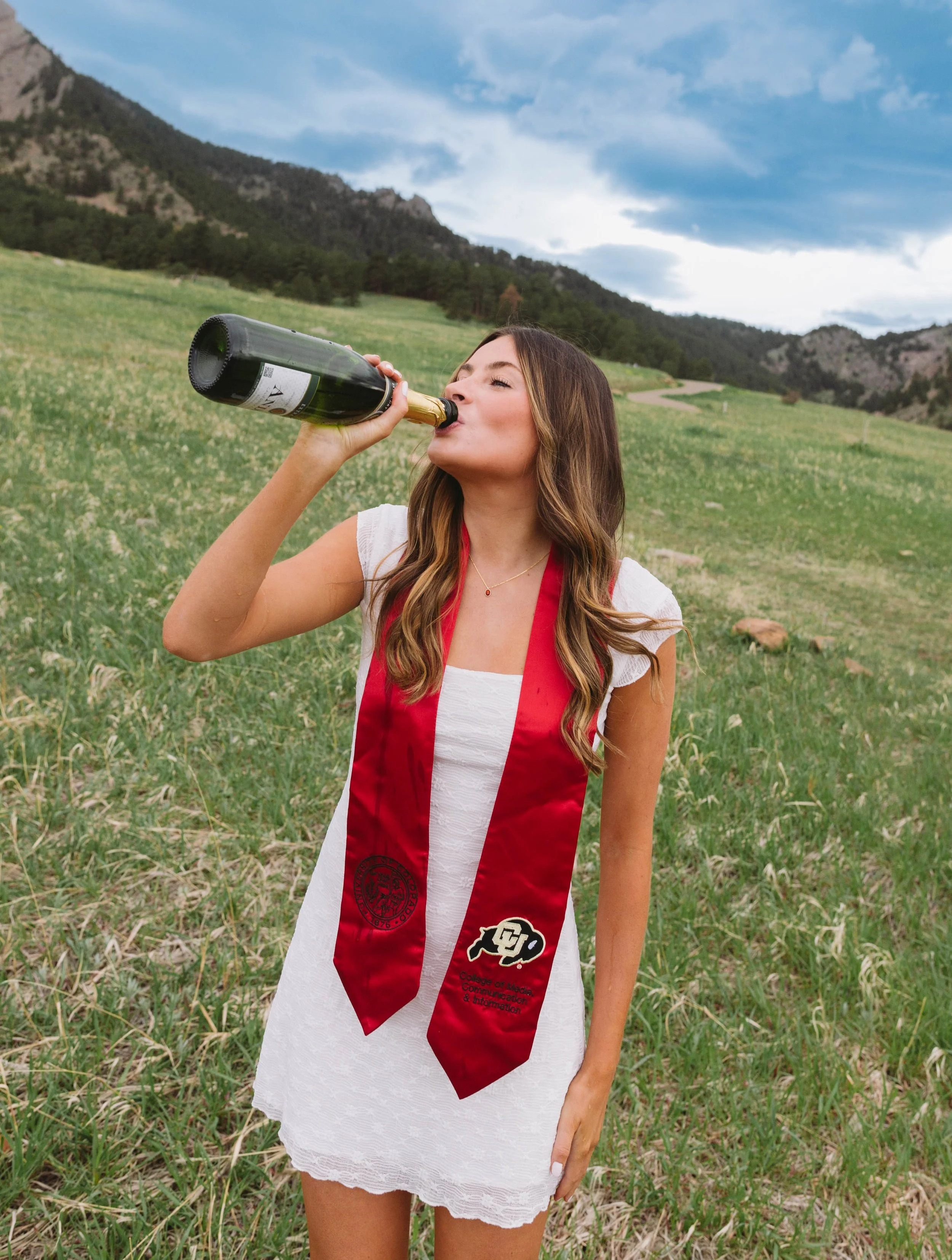 Fun CU Boulder graduation photo of a senior drinking from a champagne bottle in a field with the Flatirons behind her, capturing celebratory Boulder vibes.