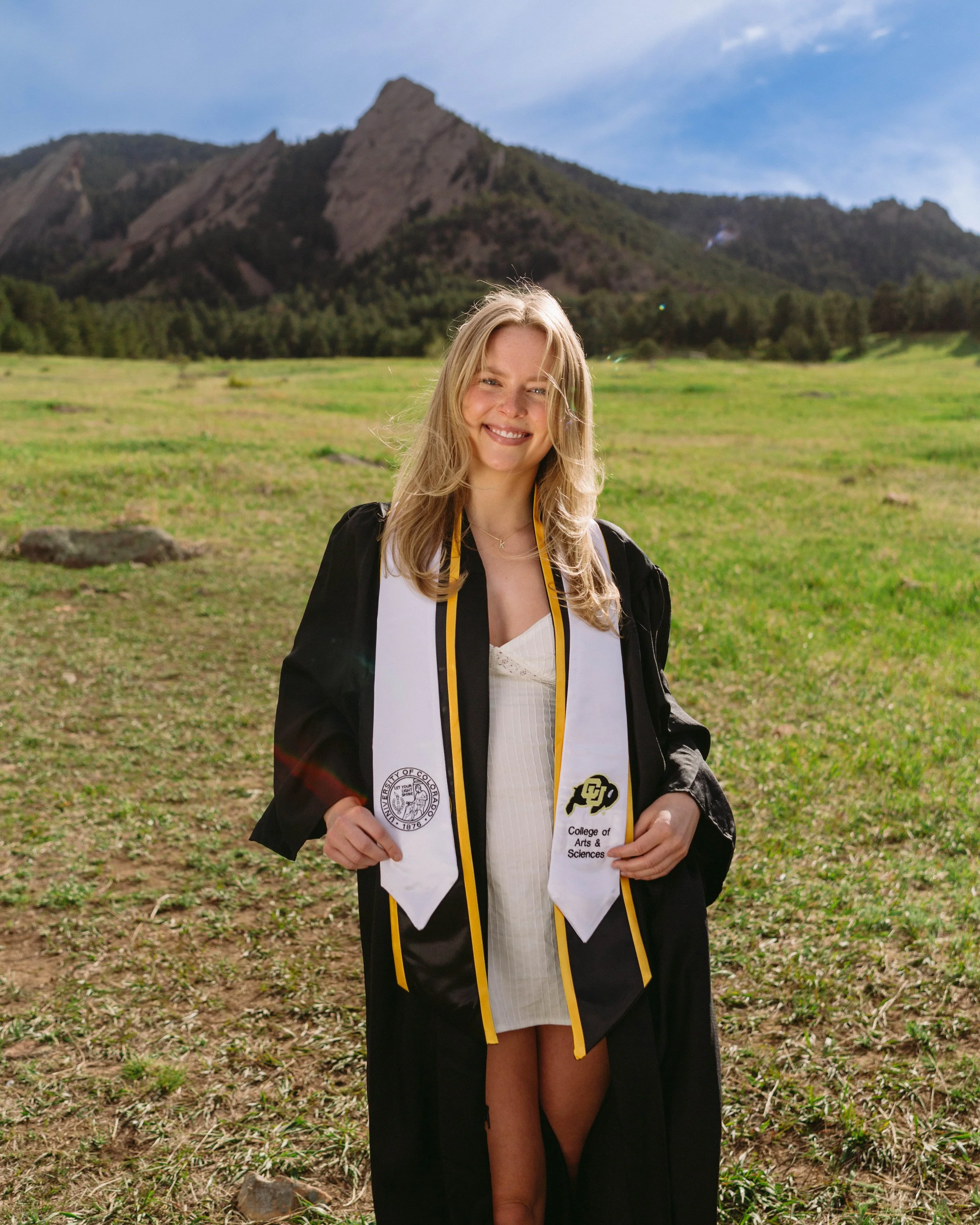 CU Boulder senior portrait featuring a graduate smiling in her cap and gown in front of the Flatirons, showcasing clean natural light and Boulder’s iconic scenery.
