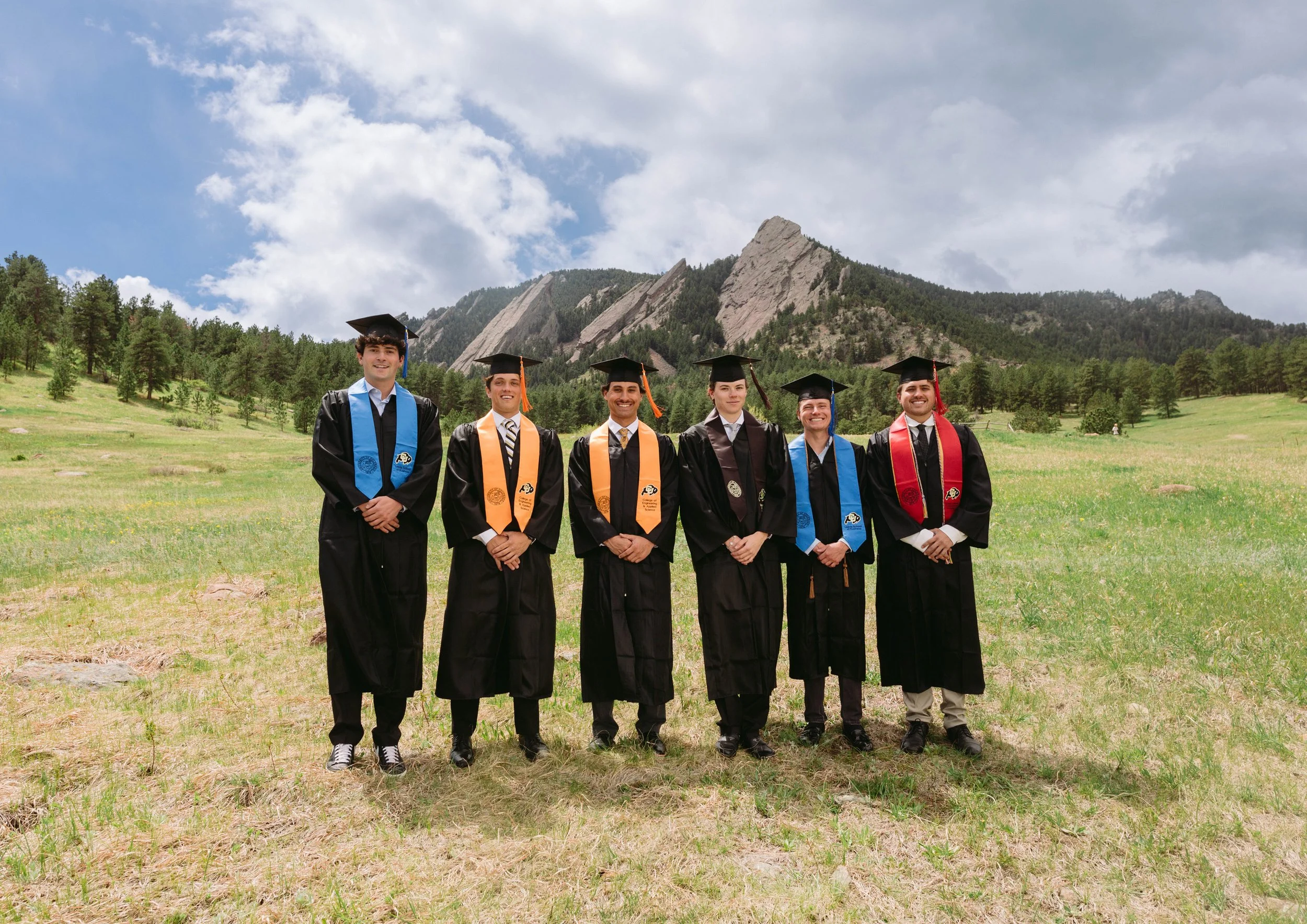 Six young men in graduation gowns and caps standing outdoors in a grassy field with trees and mountains in the background.