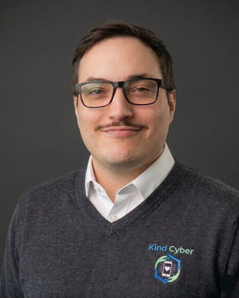 A man with dark hair and glasses, wearing a gray sweater with a logo that reads 'Kind Cyber,' smiling against a dark background.
