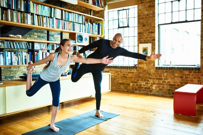 A man and a girl doing yoga together in a spacious, bright room with big windows, a brick wall, and bookshelves, both smiling and balancing on one leg.