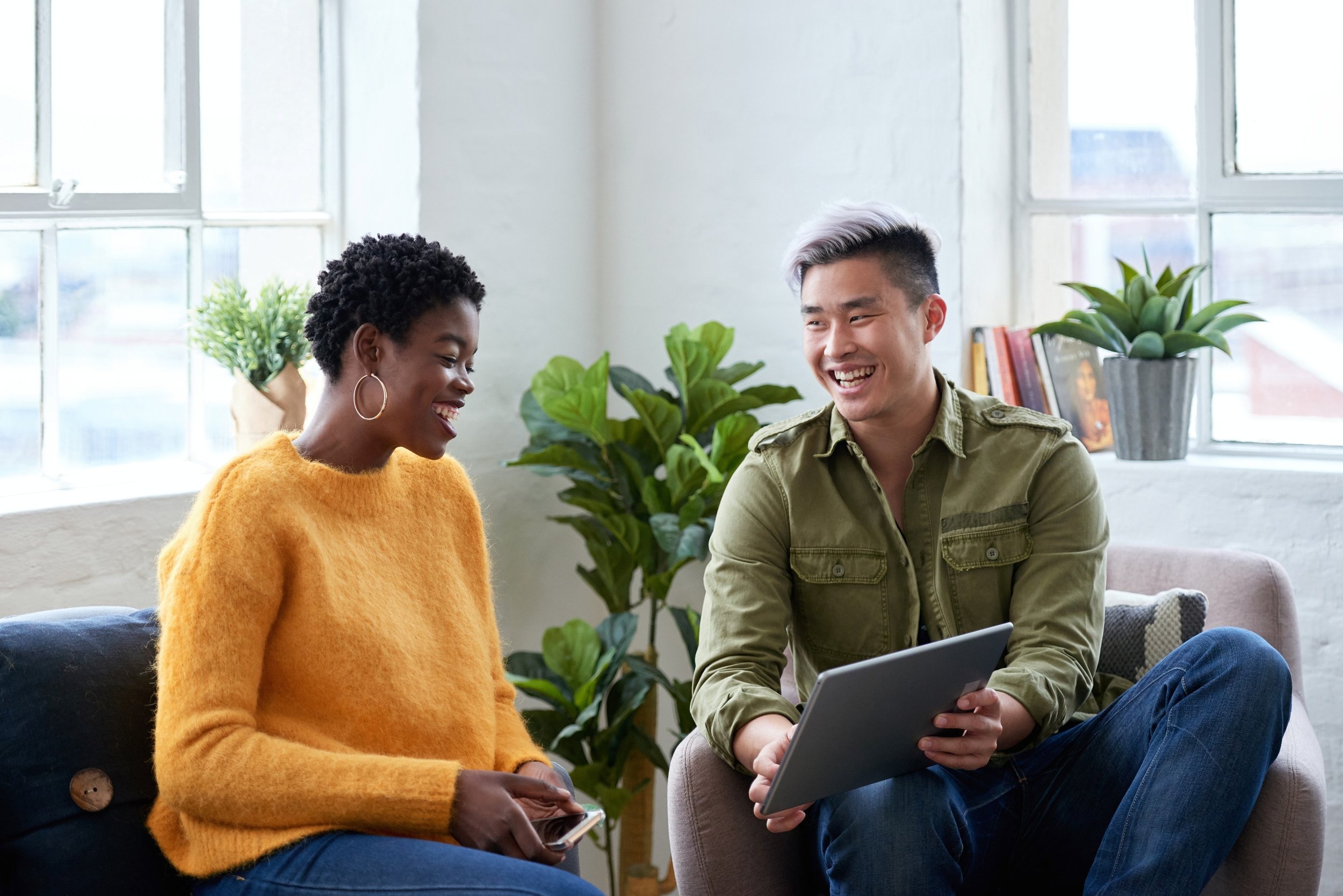A man and woman sitting on a sofa, smiling and talking, with the man holding a tablet and the woman holding a phone, in a bright room with plants.