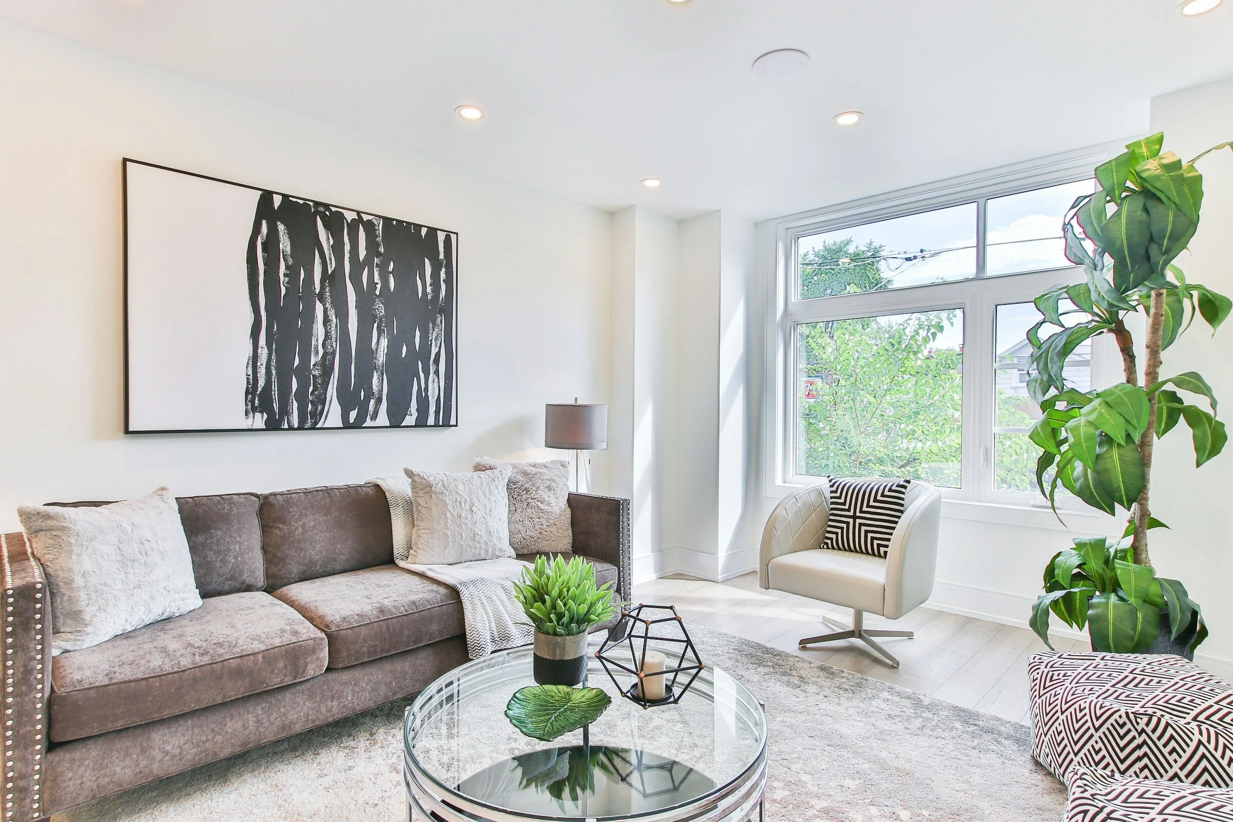 Bright living room with large window, gray sofa with plush pillows, white armchair with black and white patterned pillow, glass coffee table with green plants, black wall art, and a tall potted plant.
