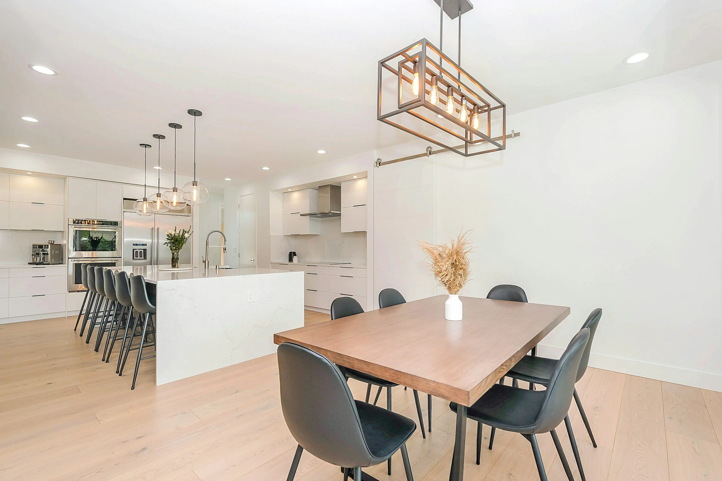 Modern kitchen and dining area with white cabinetry, a white island, and a wooden dining table surrounded by black chairs. Stylish light fixtures hang from the ceiling, and there are decorative items like a vase with dried plants on the table.