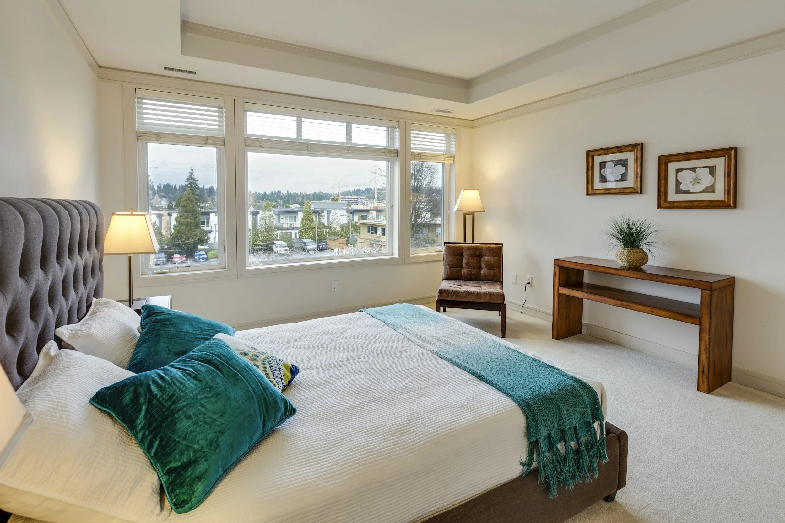 A bedroom with a bed, teal pillows, a teal blanket, a nightstand with a lamp, a brown armchair, and a wooden console table with a potted plant. Large windows show a view of neighboring buildings and trees.