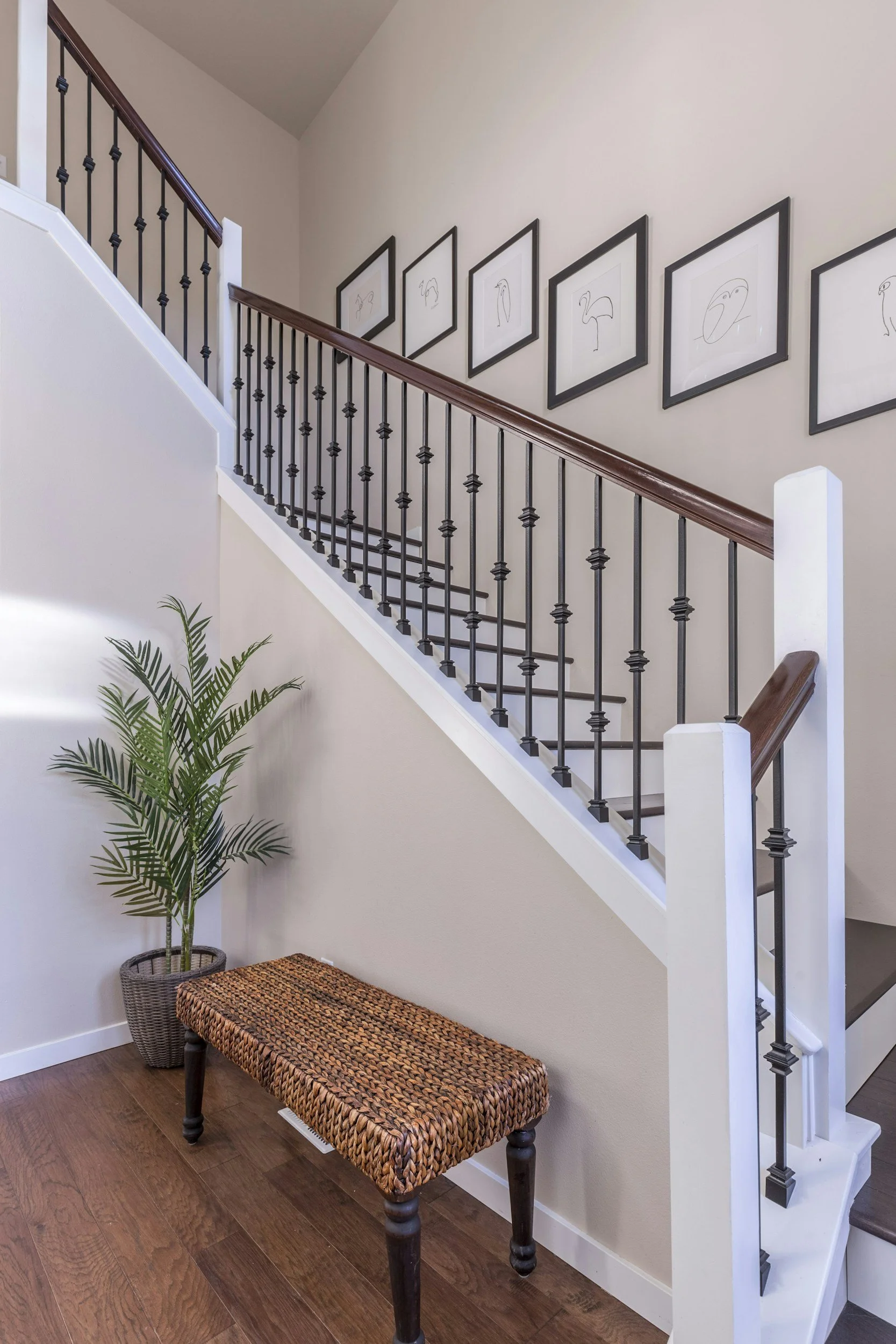 Wooden staircase with black metal railing, beige walls with framed line art photos of birds, woven bench, potted plant, and hardwood floor.