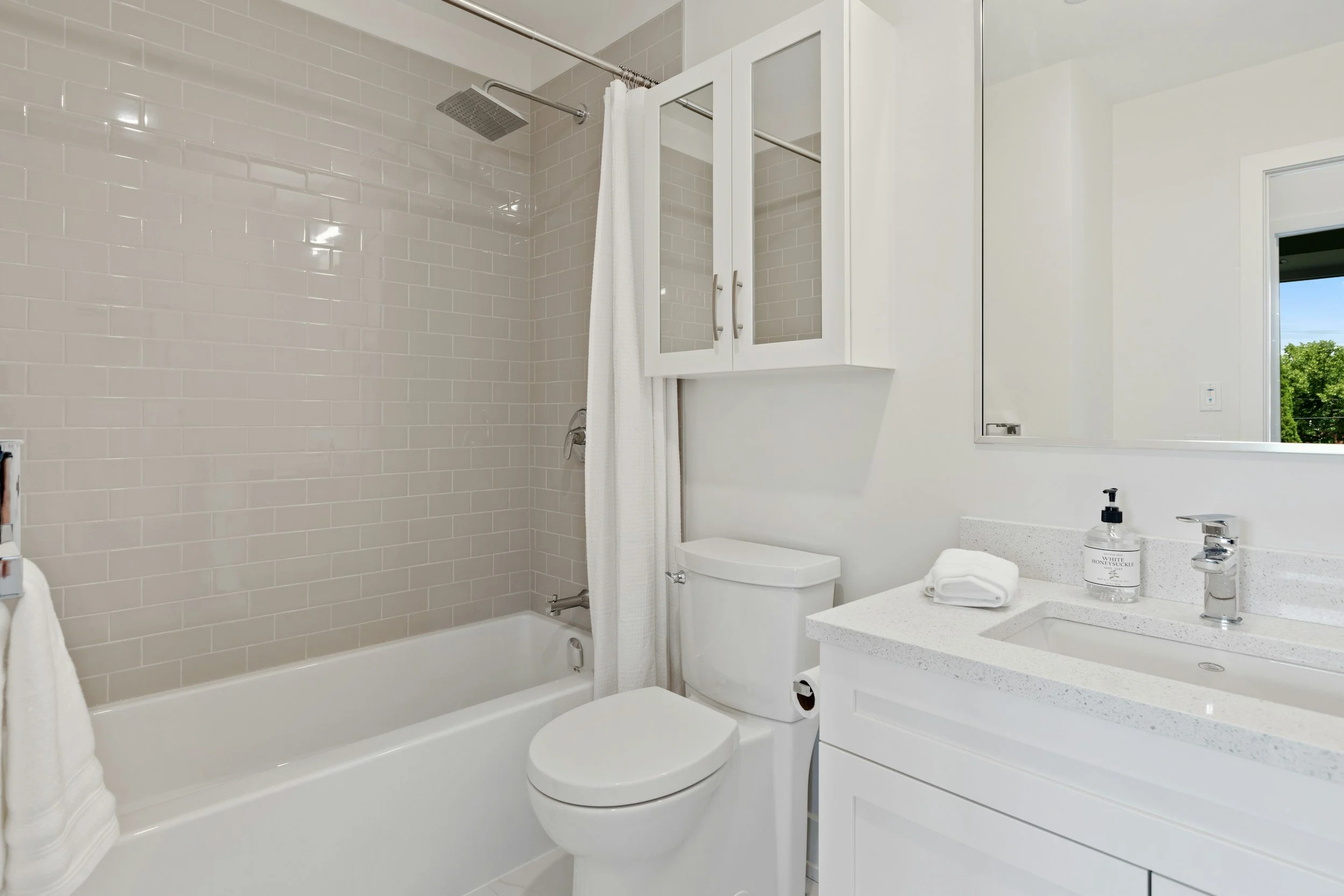 A modern bathroom featuring a bathtub with a showerhead, white subway tile walls, a white toilet, a white cabinet with mirrored doors, a white sink with a quartz countertop, and a window showing greenery outside.