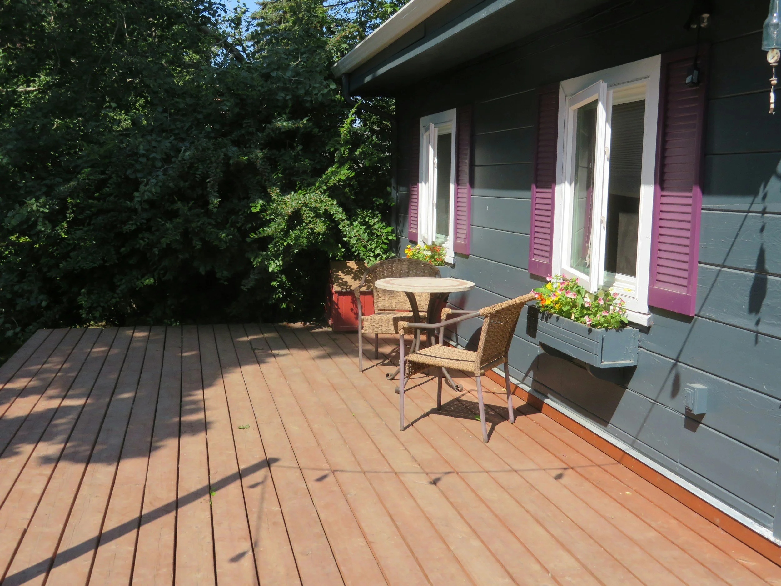 A cozy outdoor patio with a wooden deck, a small round table, two wicker chairs, colorful flower pots, and a house exterior with purple window shutters, surrounded by lush green foliage.
