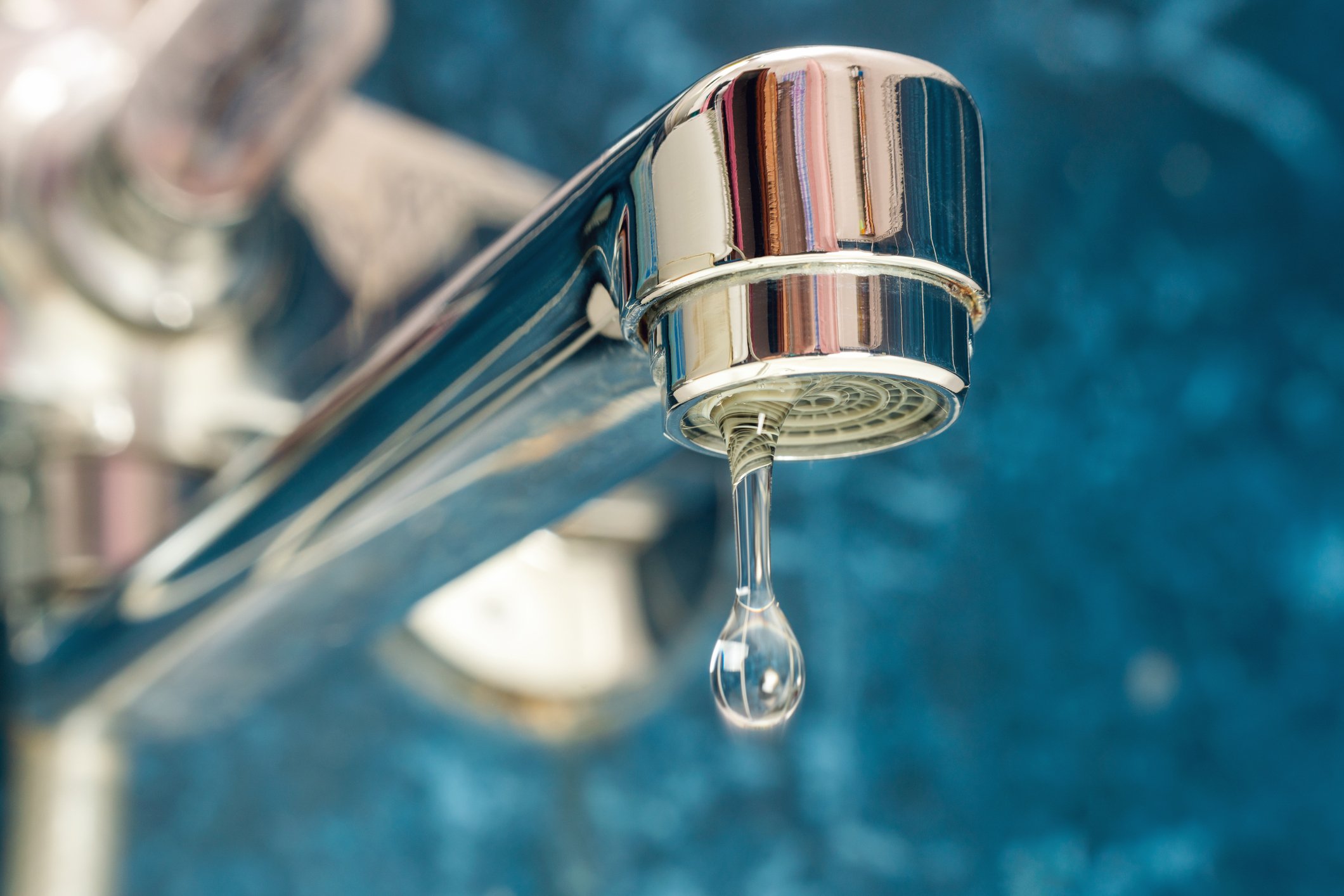 Close-up of a shiny metal faucet with water droplet hanging from the spout, set against a blurred blue background.
