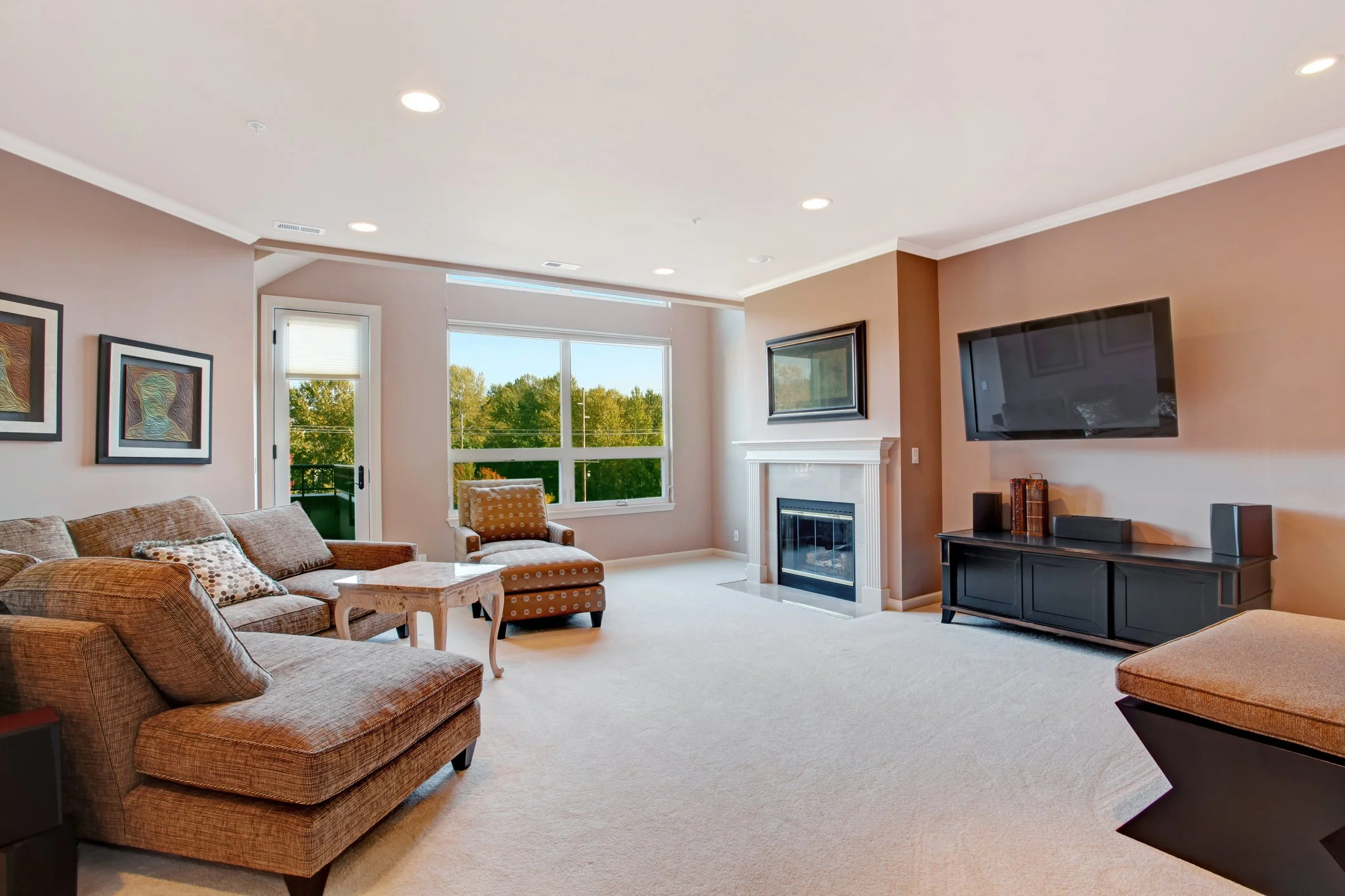 Living room with beige and brown furniture, large windows showing trees outside, a fireplace, and a wall-mounted flat-screen TV.