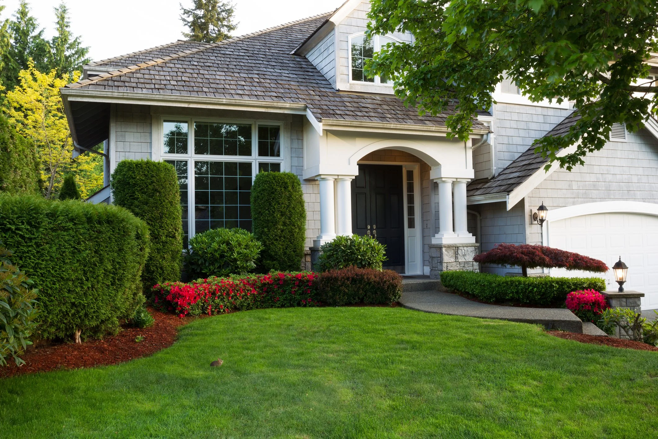 Front view of a suburban house with a well-manicured lawn, vibrant flowers, shrubs, trees, and a pathway leading to the front door.