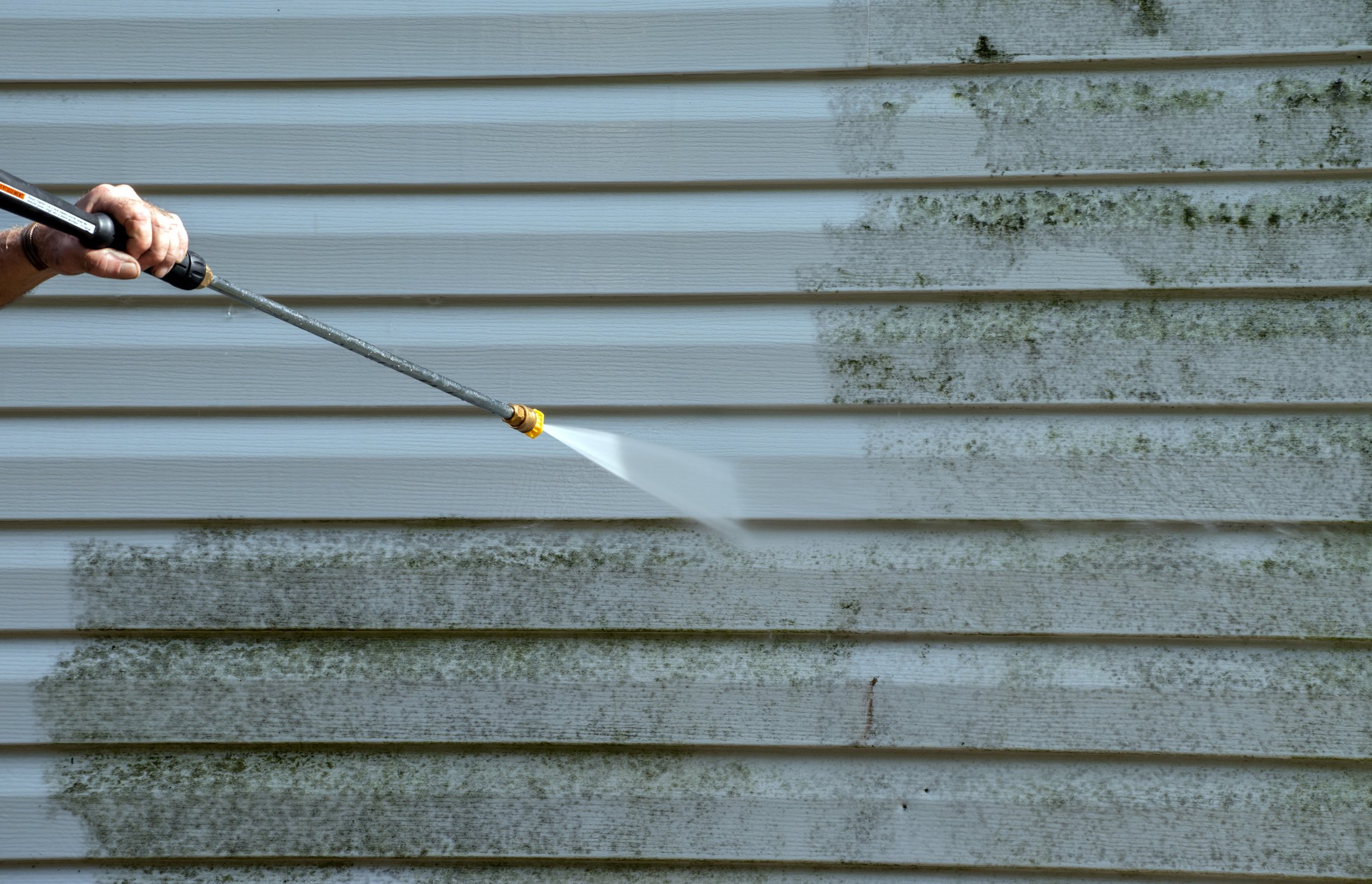 A person’s hand holding a pressure washer spray nozzle, cleaning the exterior of a white house with horizontal siding, which has dark moss or algae growth.