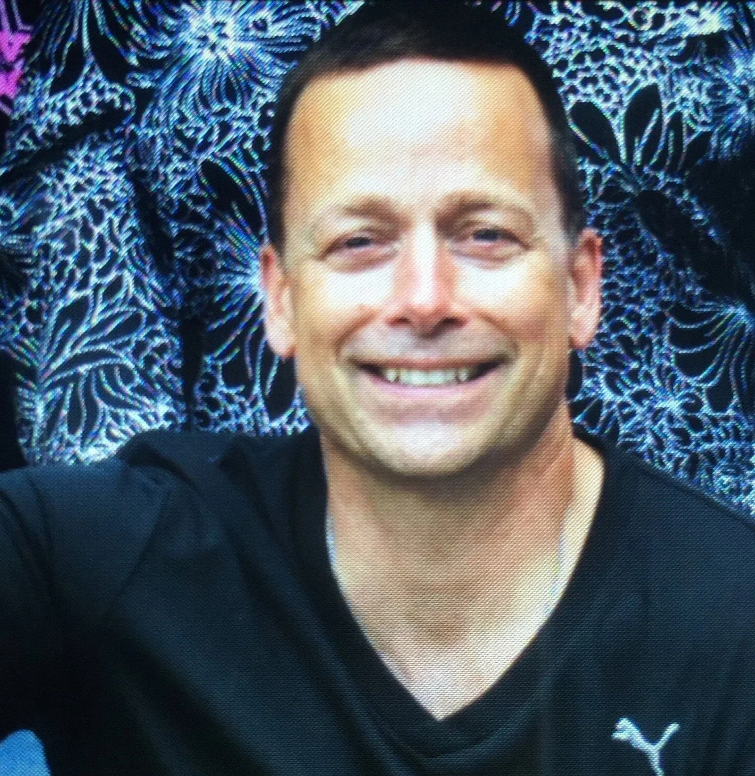 A man smiling and taking a selfie with a patterned background behind him. The background features floral and leaf designs in shades of blue, purple, and black.