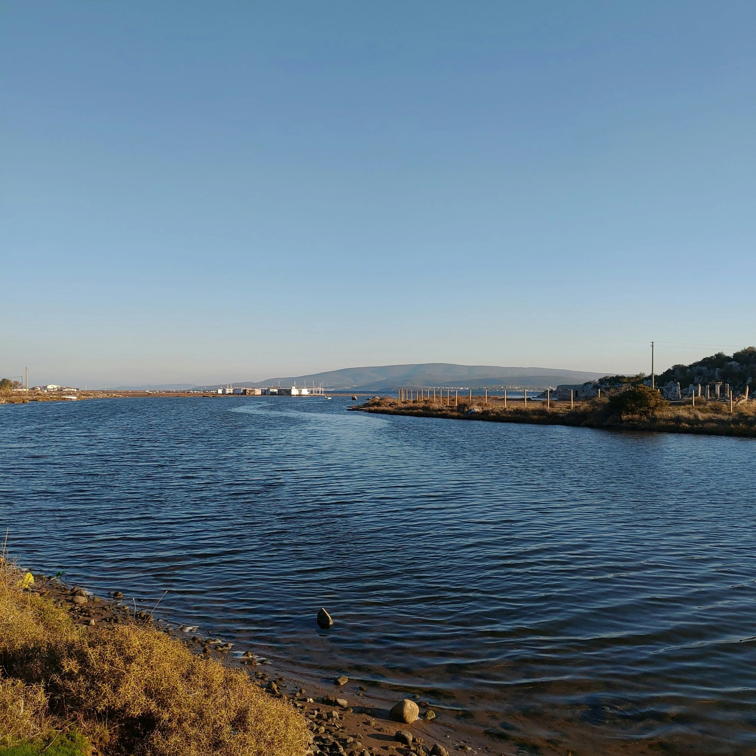 A river or lake with calm water, bordered by grassy land and small rocks, under a clear blue sky with a distant hill or mountain and a few structures in the background.