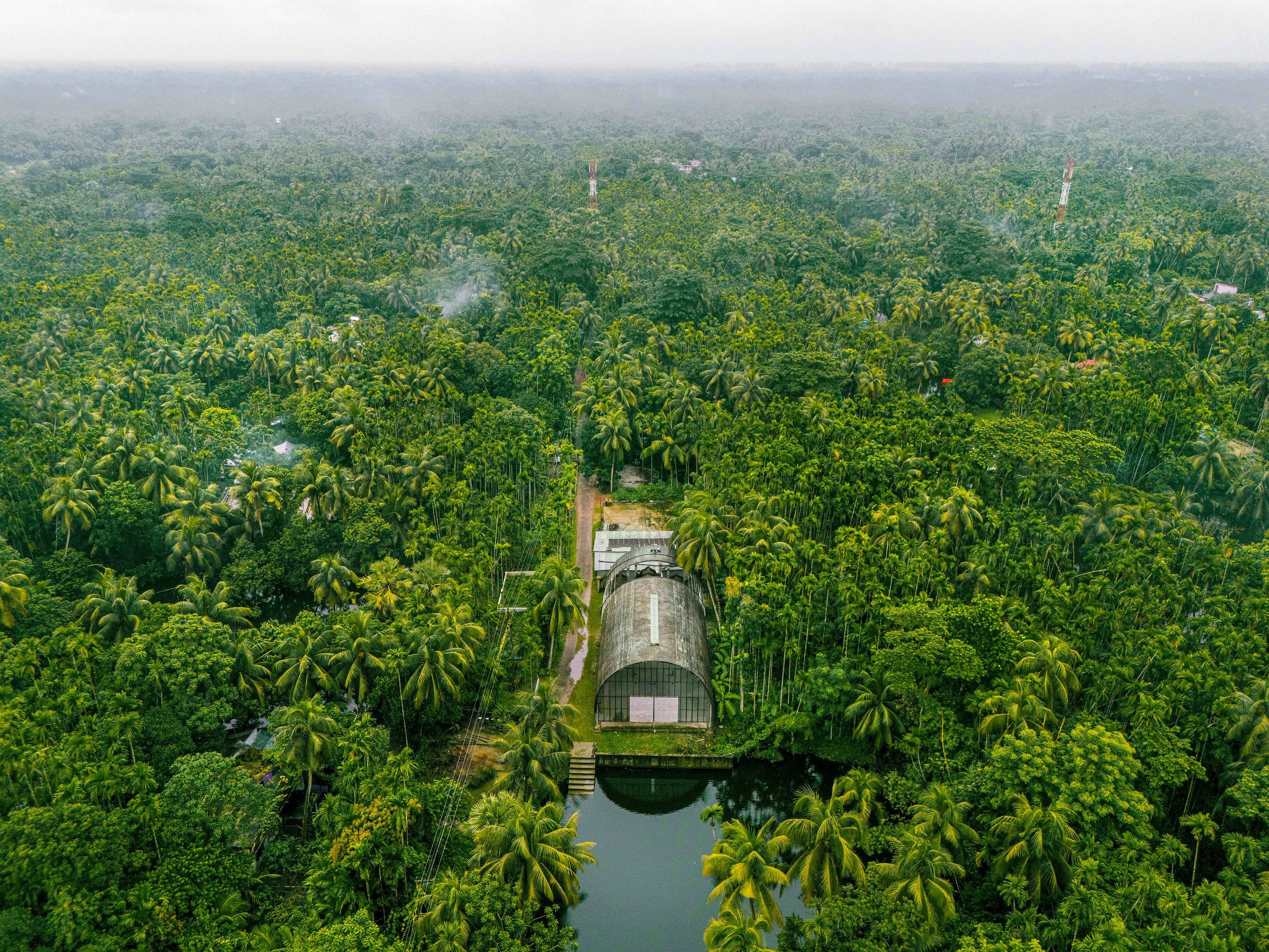 An aerial view of a lush green forest with a large greenhouse and a water body in the foreground, and communication towers in the distance.
