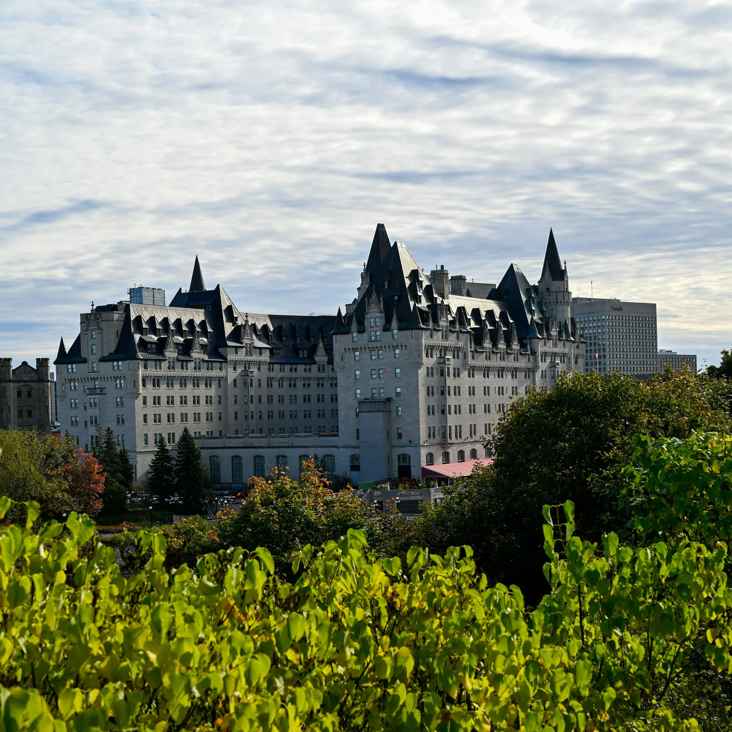 Fairy tale-style castle with pointed towers and turrets, surrounded by green trees and shrubs, under a partly cloudy sky.