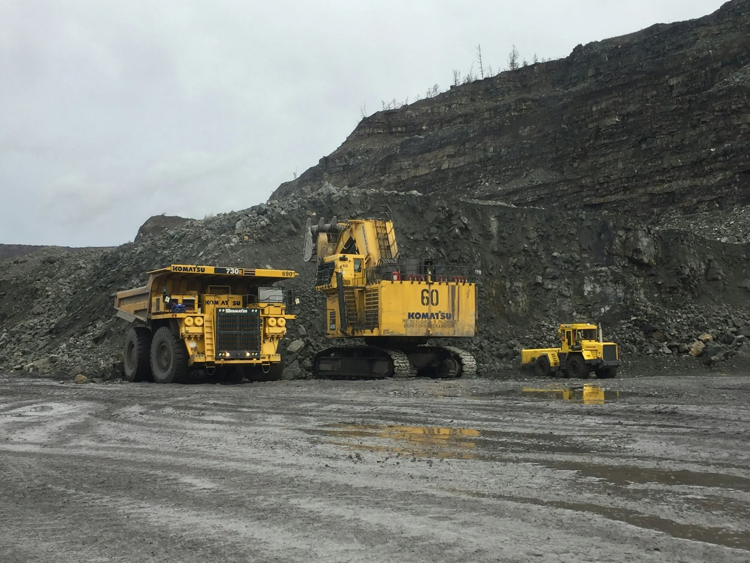 Three large construction vehicles, including a dump truck, an excavator, and a small wheel loader, are parked on a muddy construction site in front of a rocky hill.