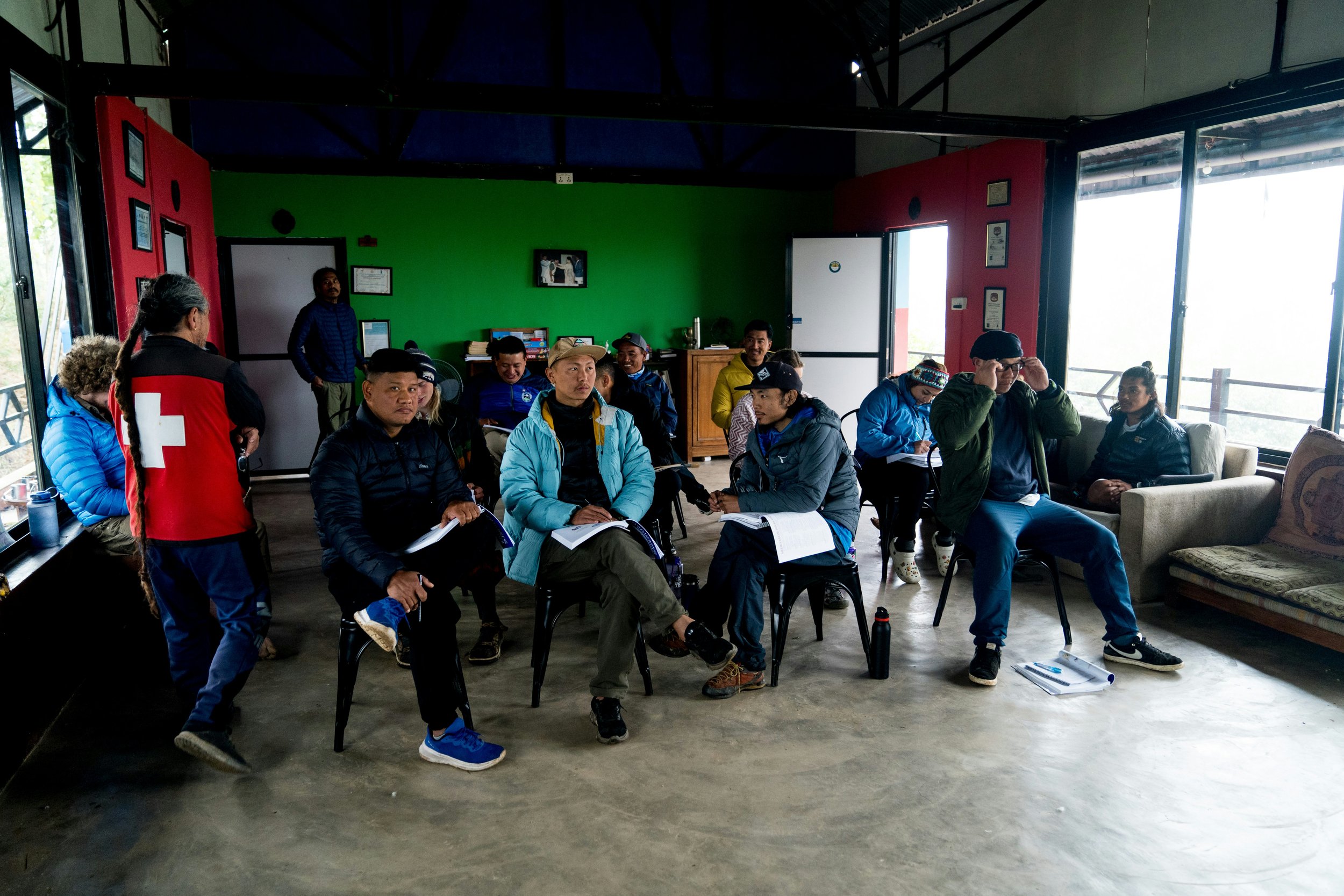 Group of people gathered inside a colorful room, sitting on chairs and couches, some are taking notes, with a woman wearing a red jacket with a white cross on the back standing on the left.