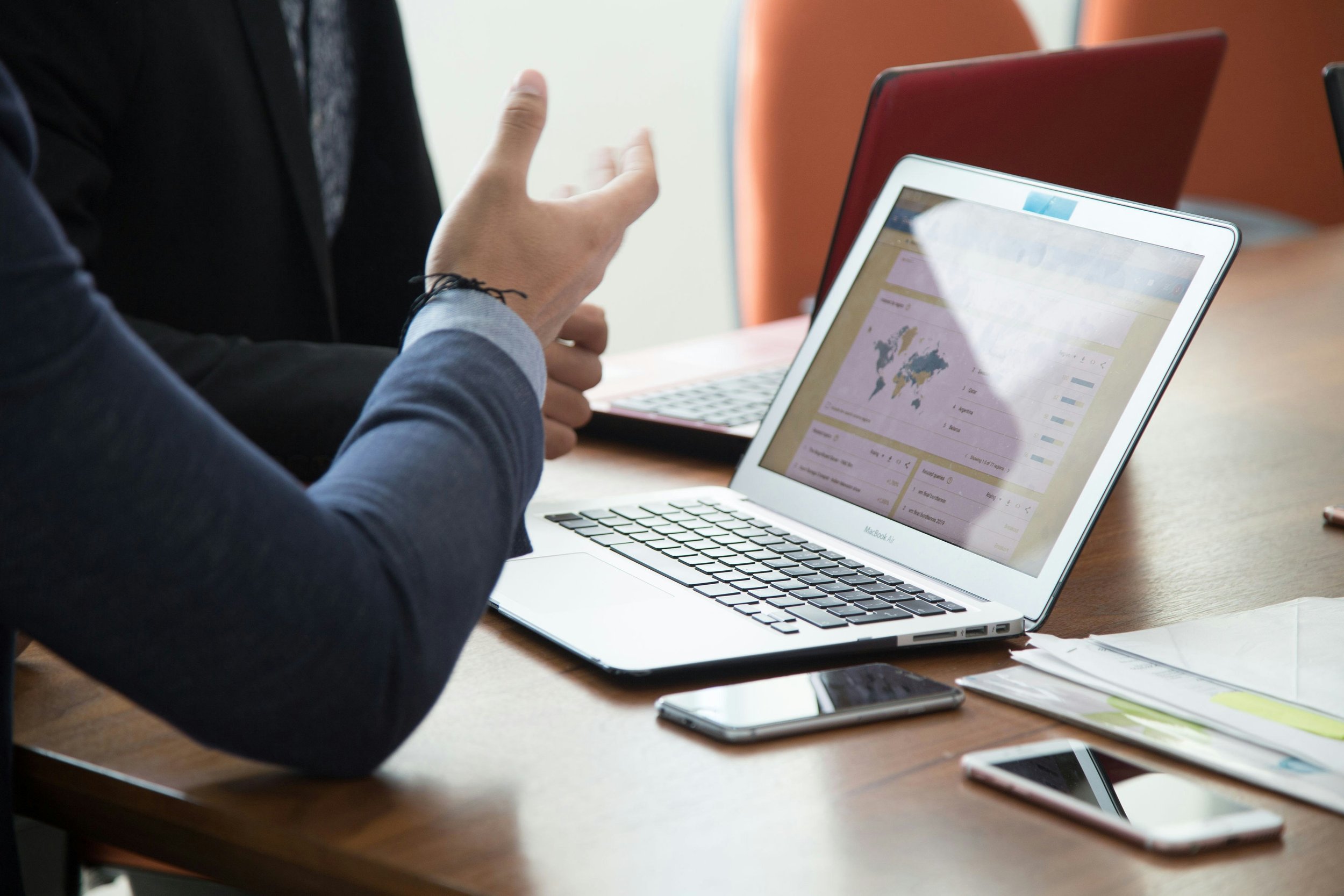 Person in a suit gesturing while discussing data on a laptop in a meeting room with other devices and documents on the table.