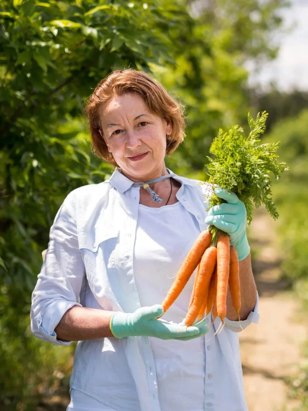 A woman holding fresh carrots and leafy greens in a garden with lush green foliage.