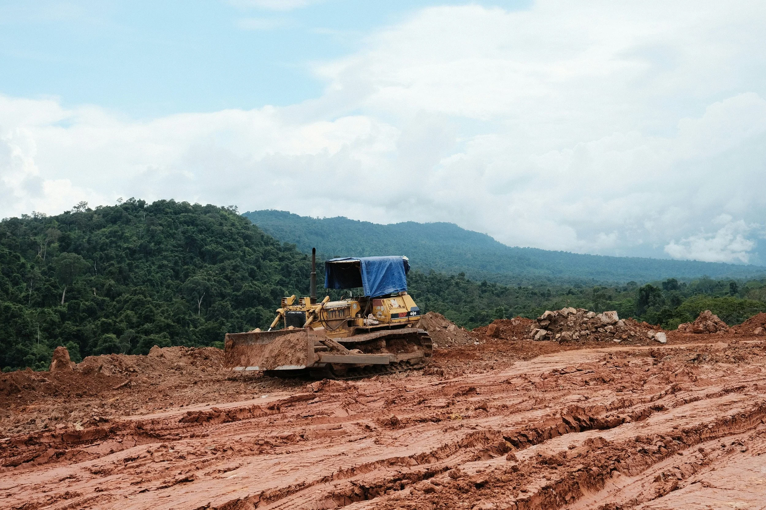 A bulldozer working on a dirt construction site in a rural area with green hills and mountains in the background.