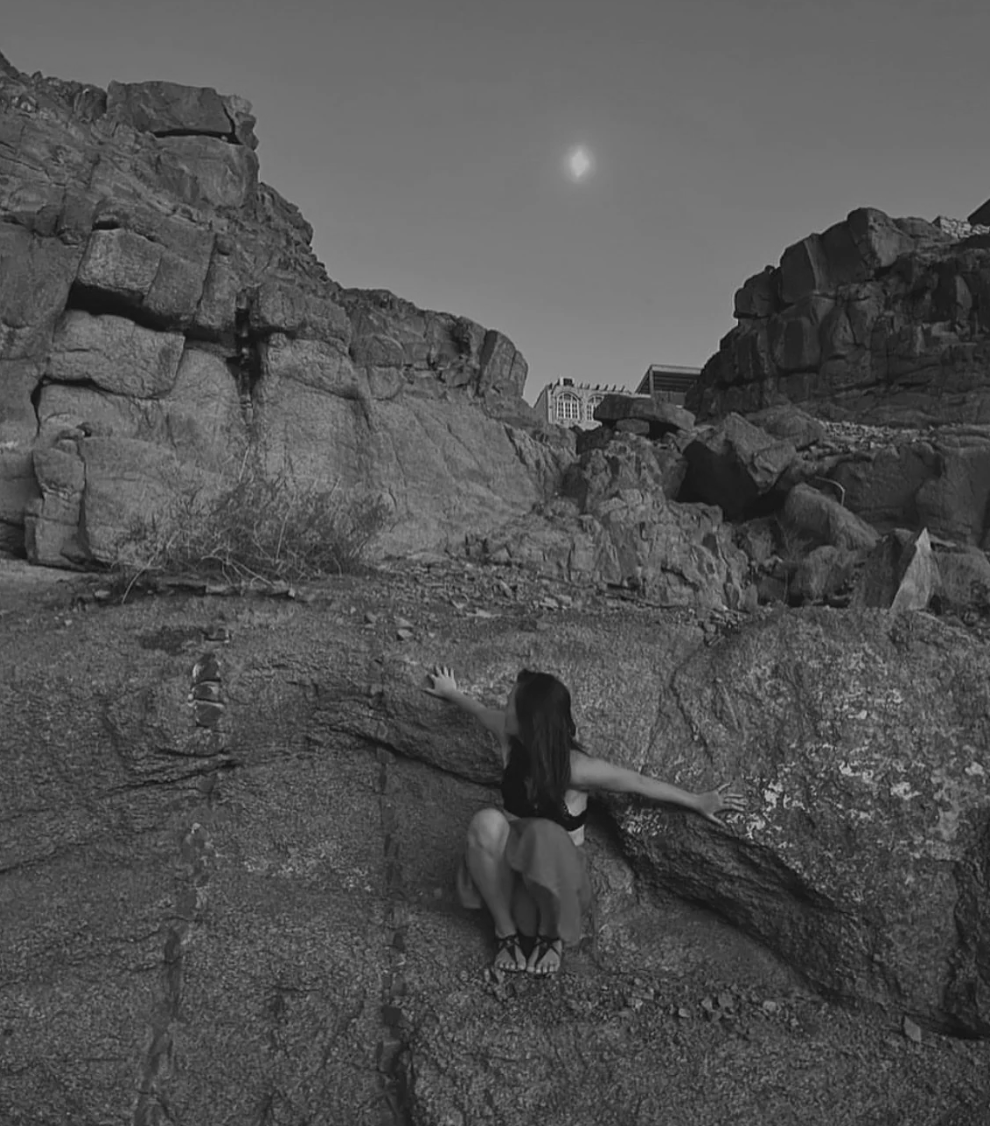 A woman with long dark hair sitting on a large rock formation in a desert landscape with large boulders and cliffs, under a clear sky with the moon visible.