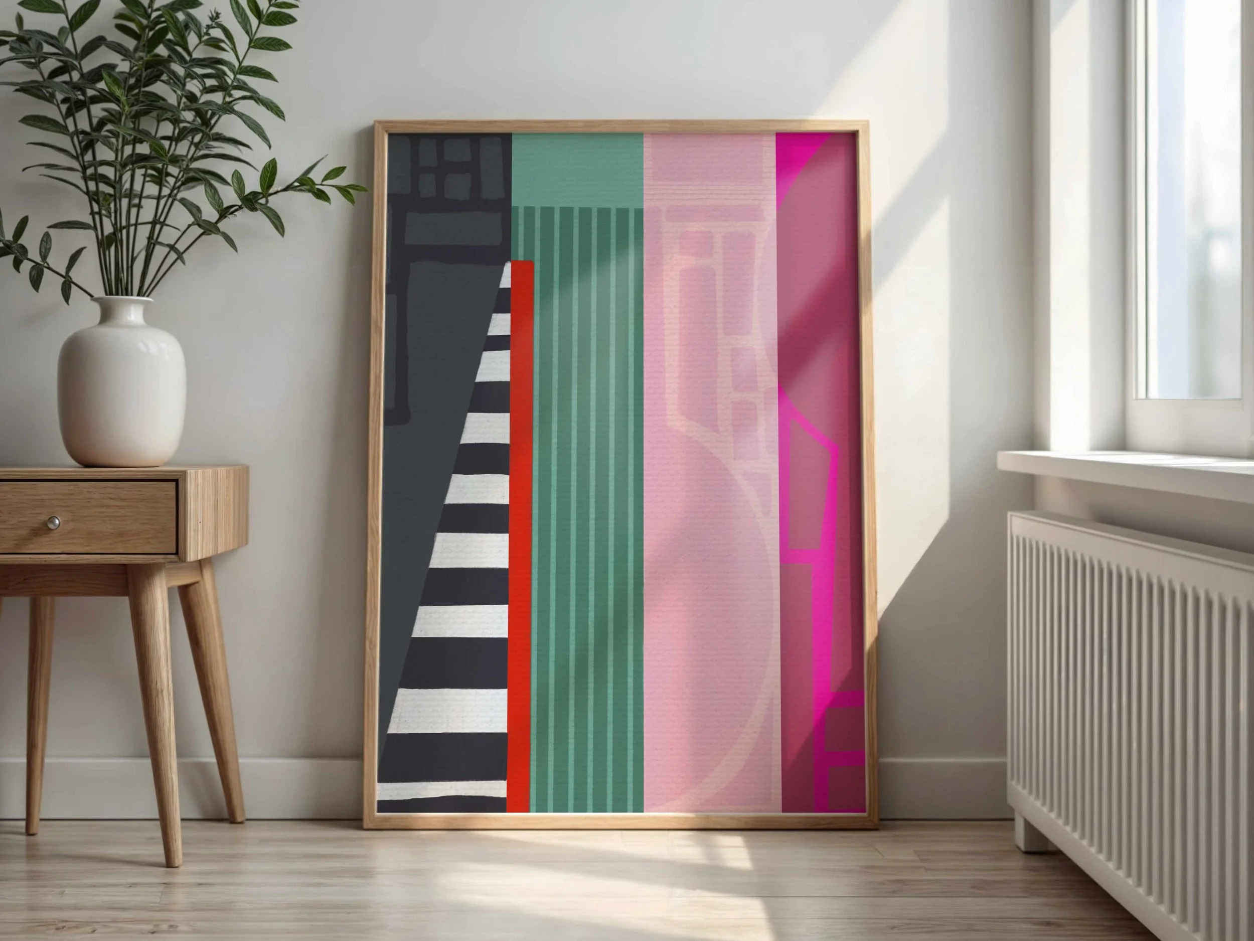 Colorful abstract artwork in a wooden frame leaning against a wall, next to a wooden side table with a white vase and green plant, in a bright room with a window.