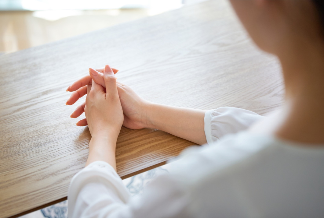 Person with clasped hands resting on a wooden table, seen from behind.