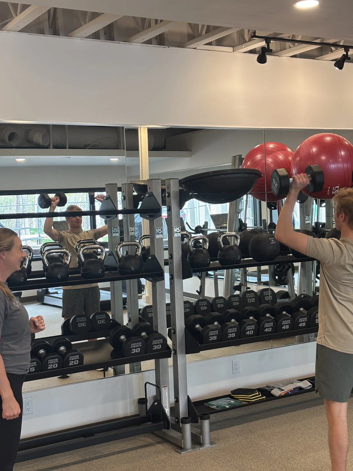 A woman in black workout leggings is lifting a barbell in a gym with other people exercising in the background.