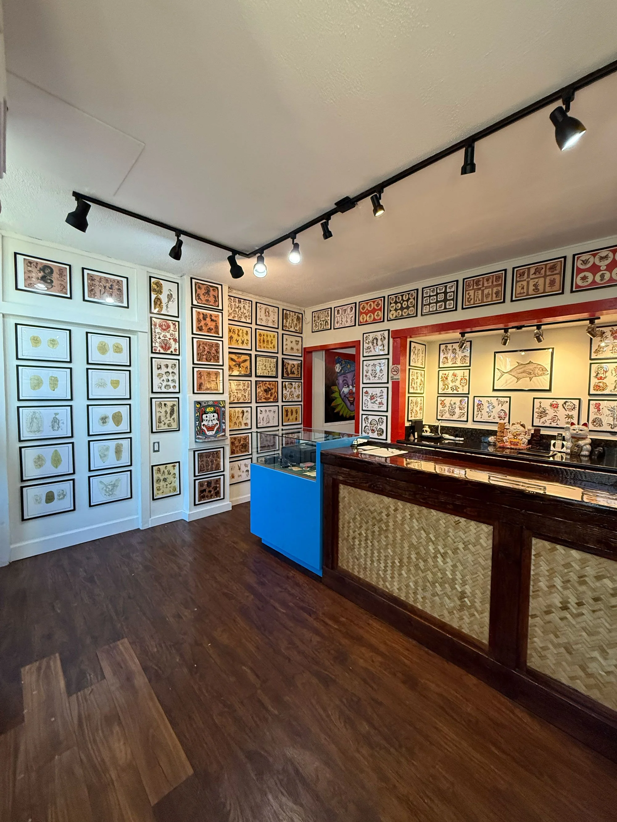 Interior of a tattoo shop with framed tattoo flash art on the walls, a black and wood reception counter, and a display case with more artwork.