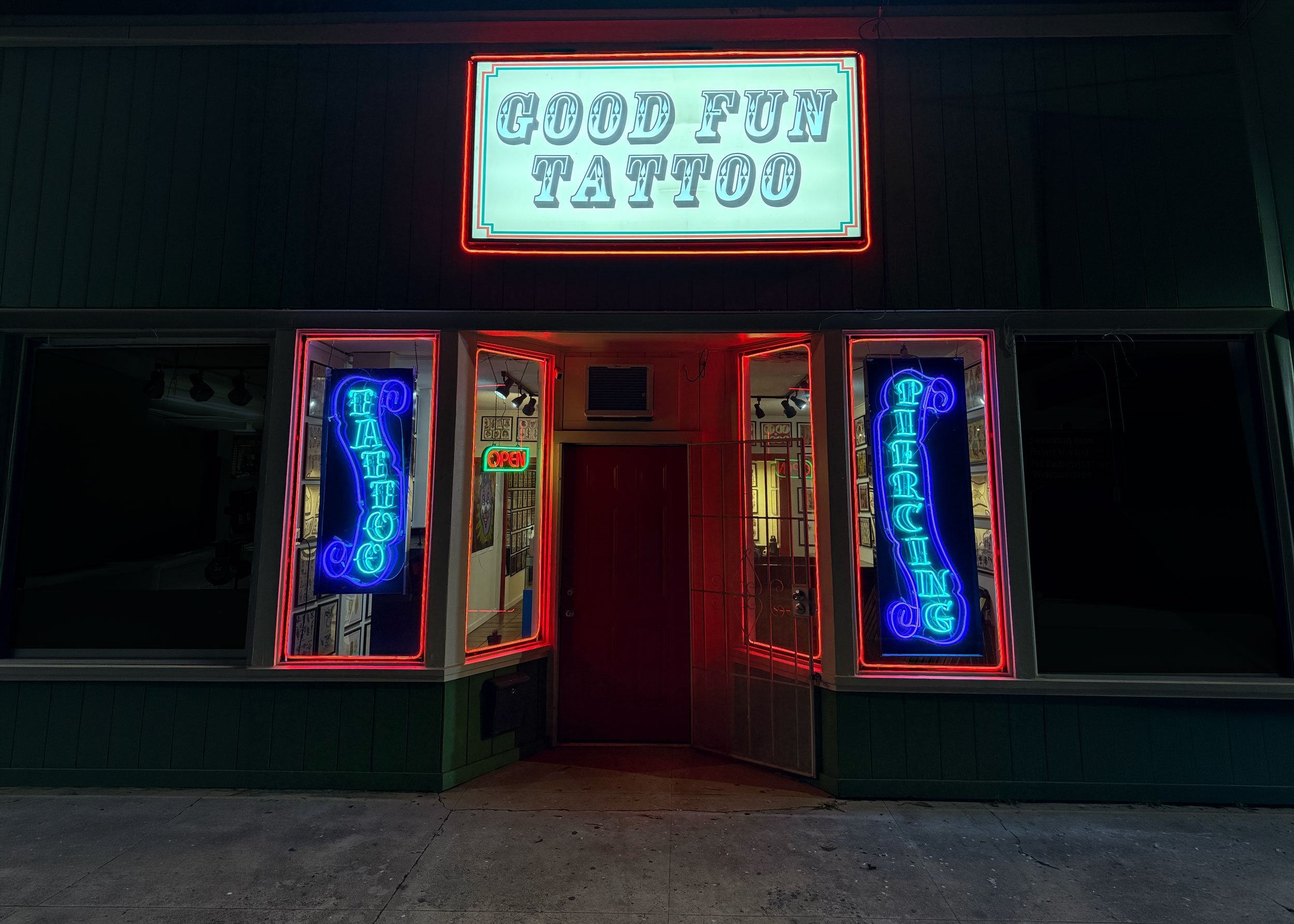 Neon sign outside tattoo shop reading 'Good Fun Tattoo' with neon signs for 'Tattoo' and 'Piercing' on each side of the entrance, and a small 'Open' sign inside near the door.