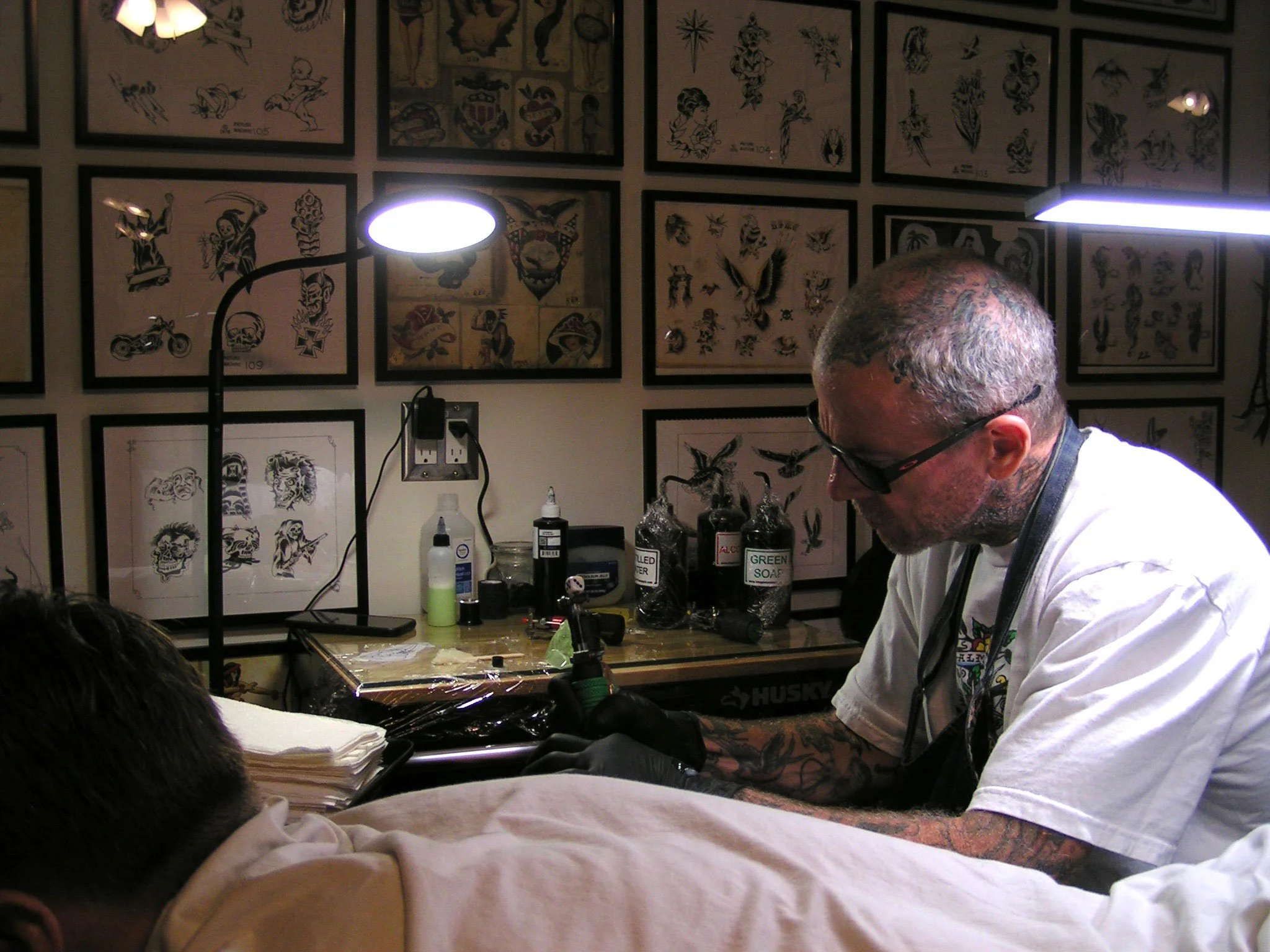 Tattoo artist working on a client in a tattoo studio, with tattoo artwork displayed on the walls behind.