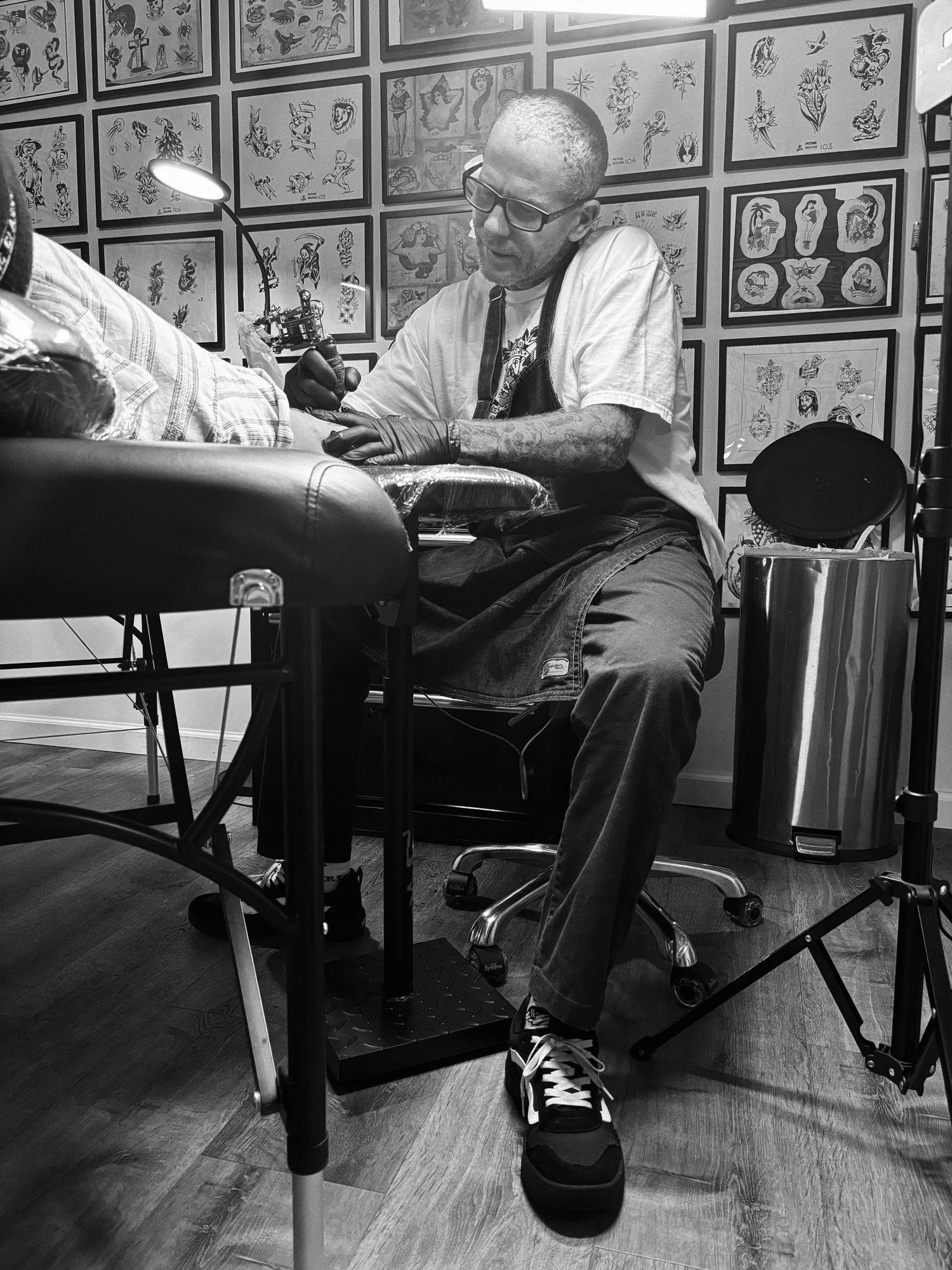 Tattoo artist working on a client's arm in a tattoo studio, with tattoo designs displayed on the walls in black and white.