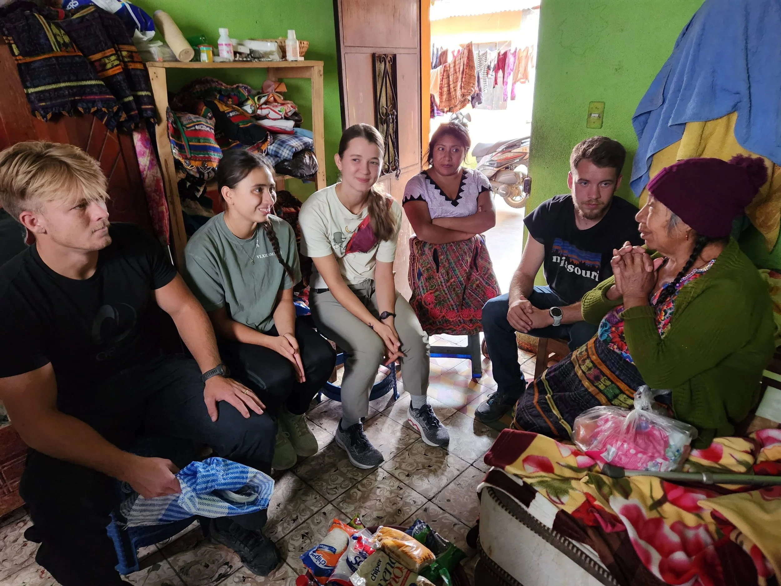 Group of six people sitting and talking inside a small room with green walls, cluttered shelves, and an open door leading outside where clothes hang to dry.
