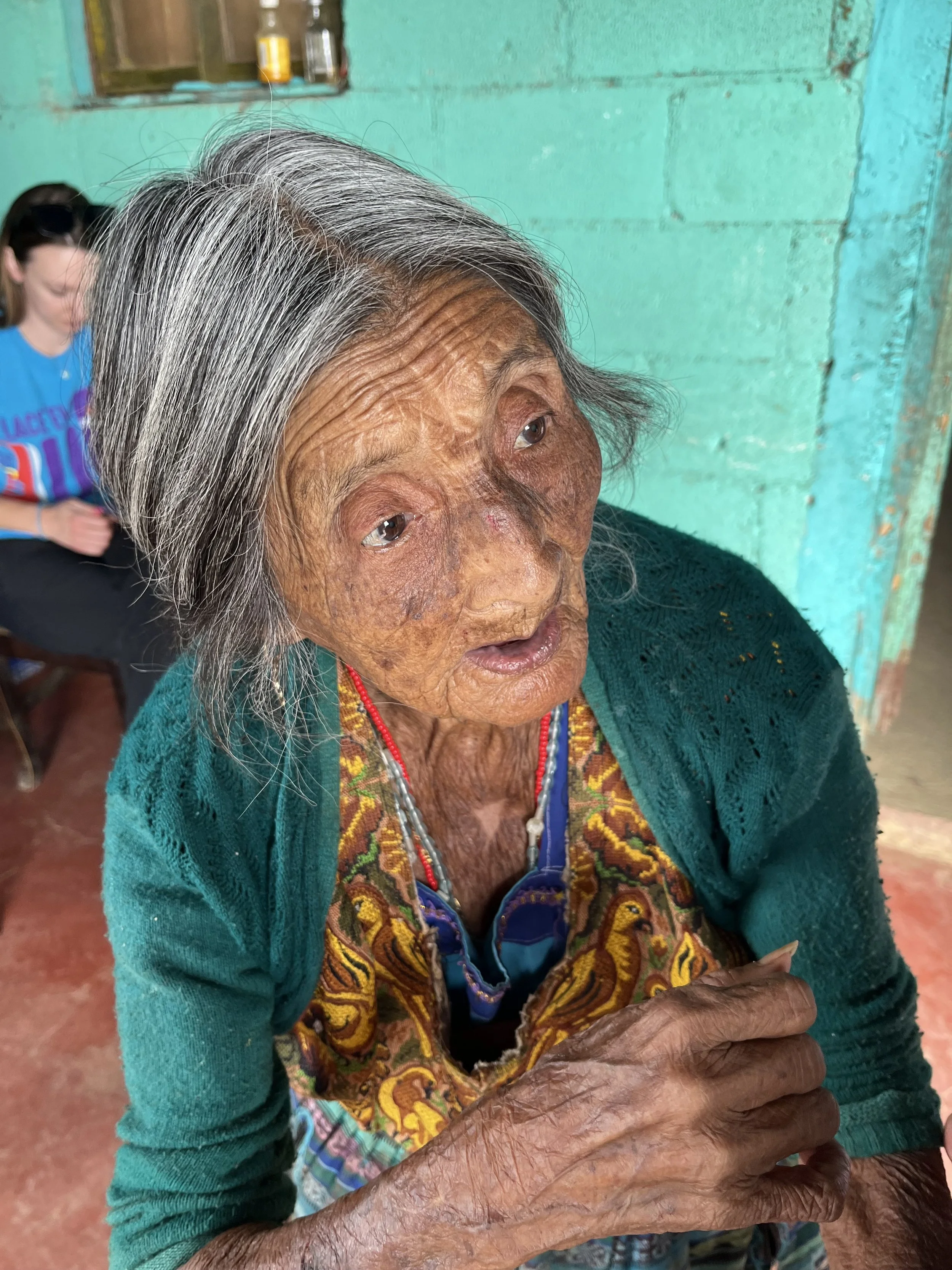 An elderly woman with gray hair and deep skin wrinkles, wearing a colorful embroidered vest and a green cardigan, sitting indoors against a turquoise wall.