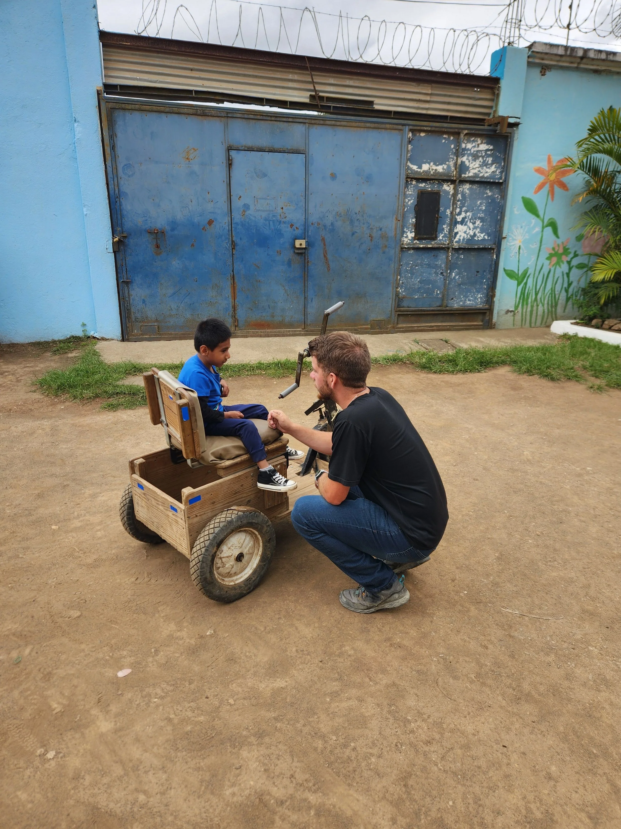 A man kneeling in front of a young boy sitting in a wooden wheelchair outdoors, with a blue wall and decorated mural in the background.