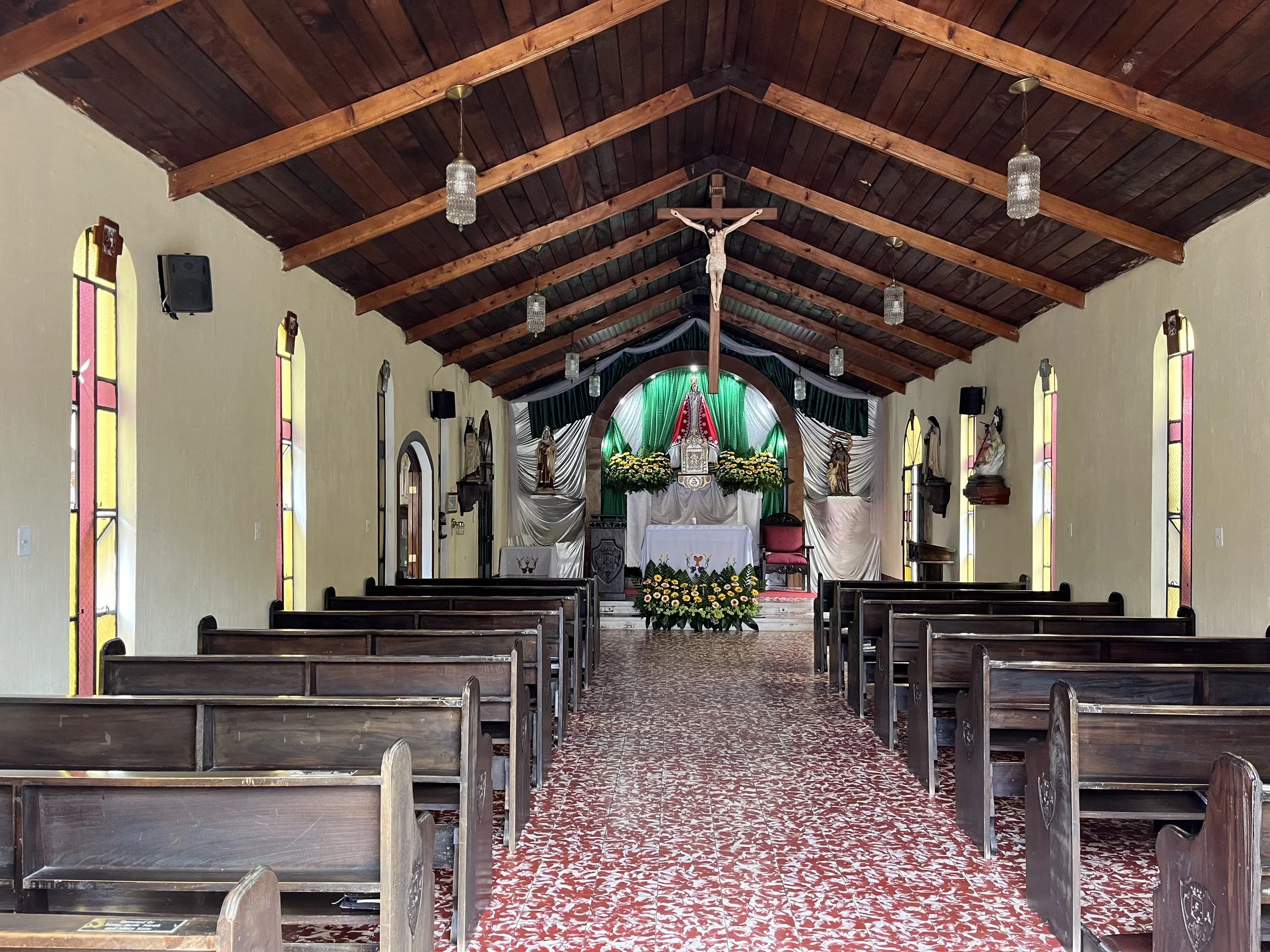 Interior of a church with wooden pews, stained glass windows, a cross hanging from the ceiling, and an altar decorated with flowers and religious statues.