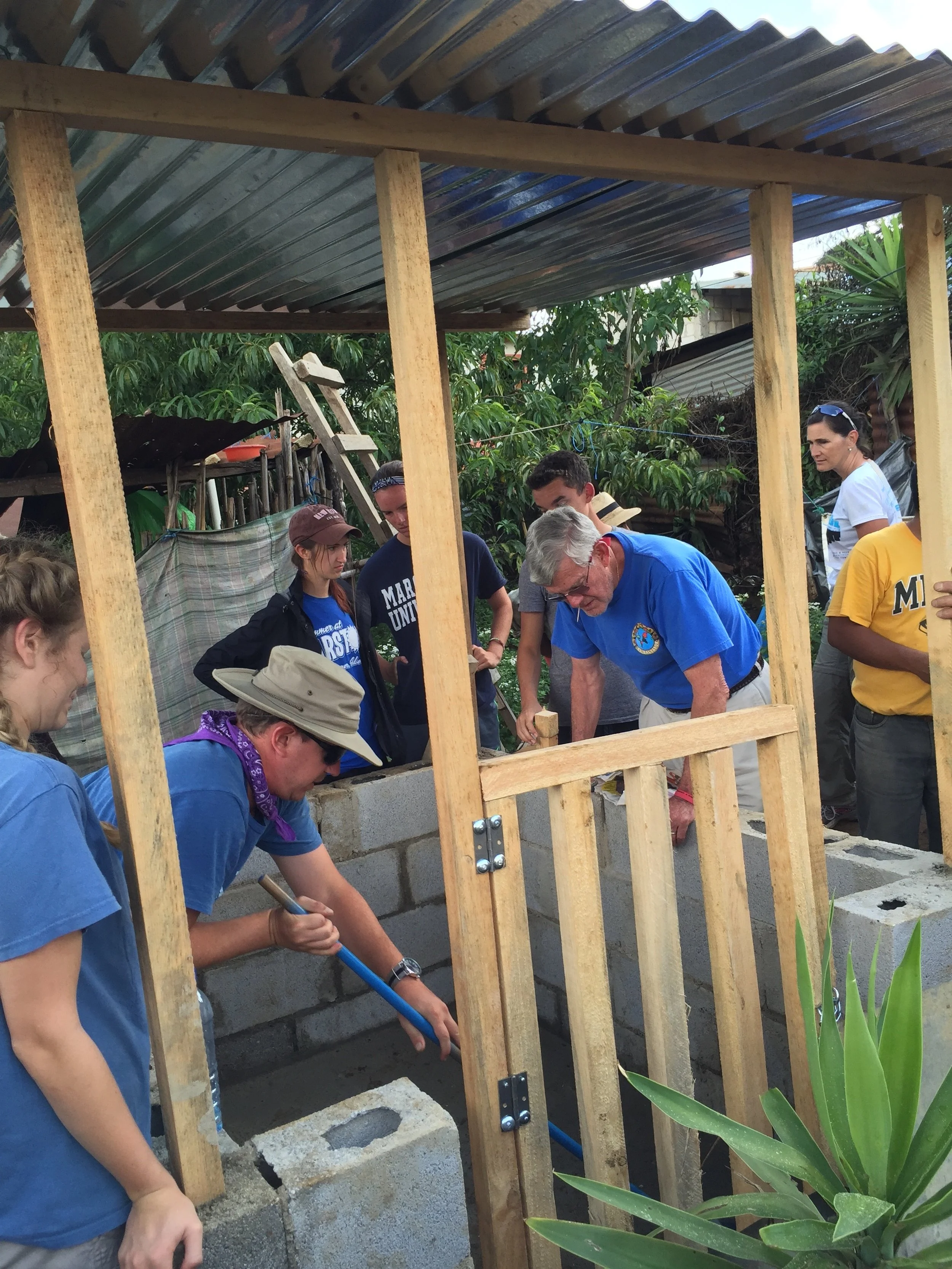 Group of people building a small structure with wooden beams and concrete blocks outdoors, surrounded by greenery.