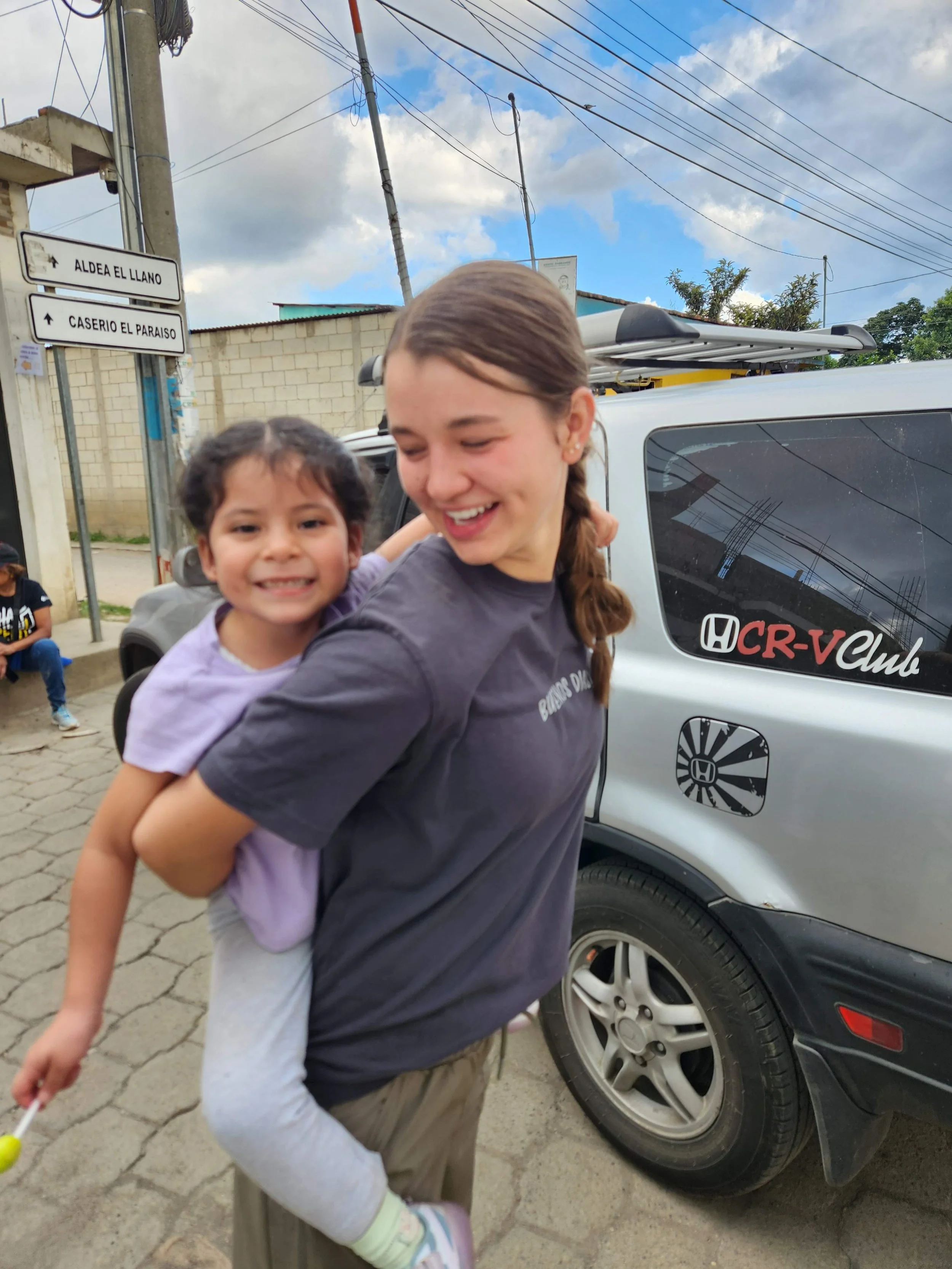 A smiling young woman carrying a girl on her back in a street scene, with a car and street signs in the background.
