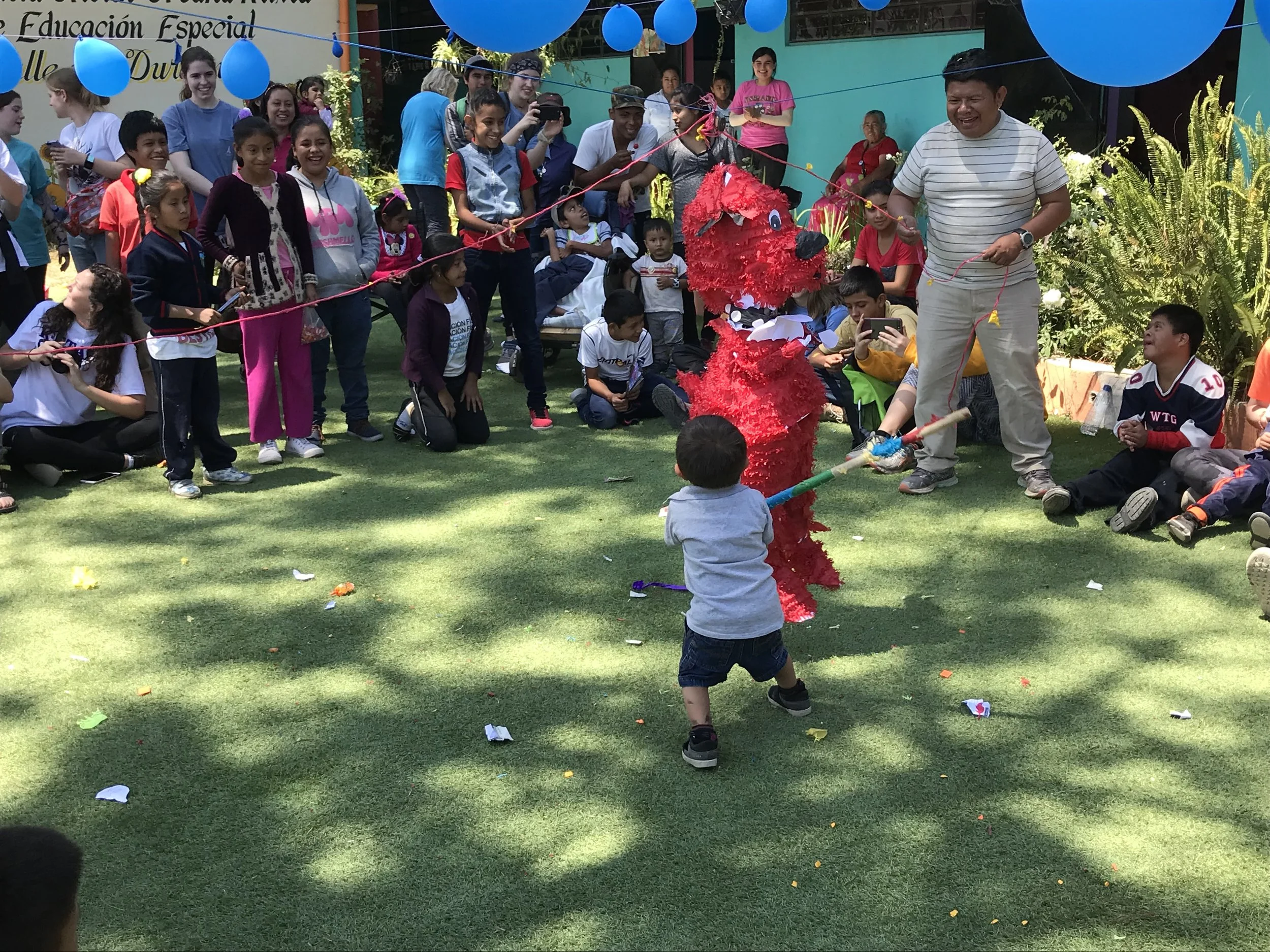 Children and adults at a celebration, with a red piñata shaped like an animal in the center, as a young boy prepares to hit it with a stick during daytime. Bright blue balloons and colorful decorations are visible.