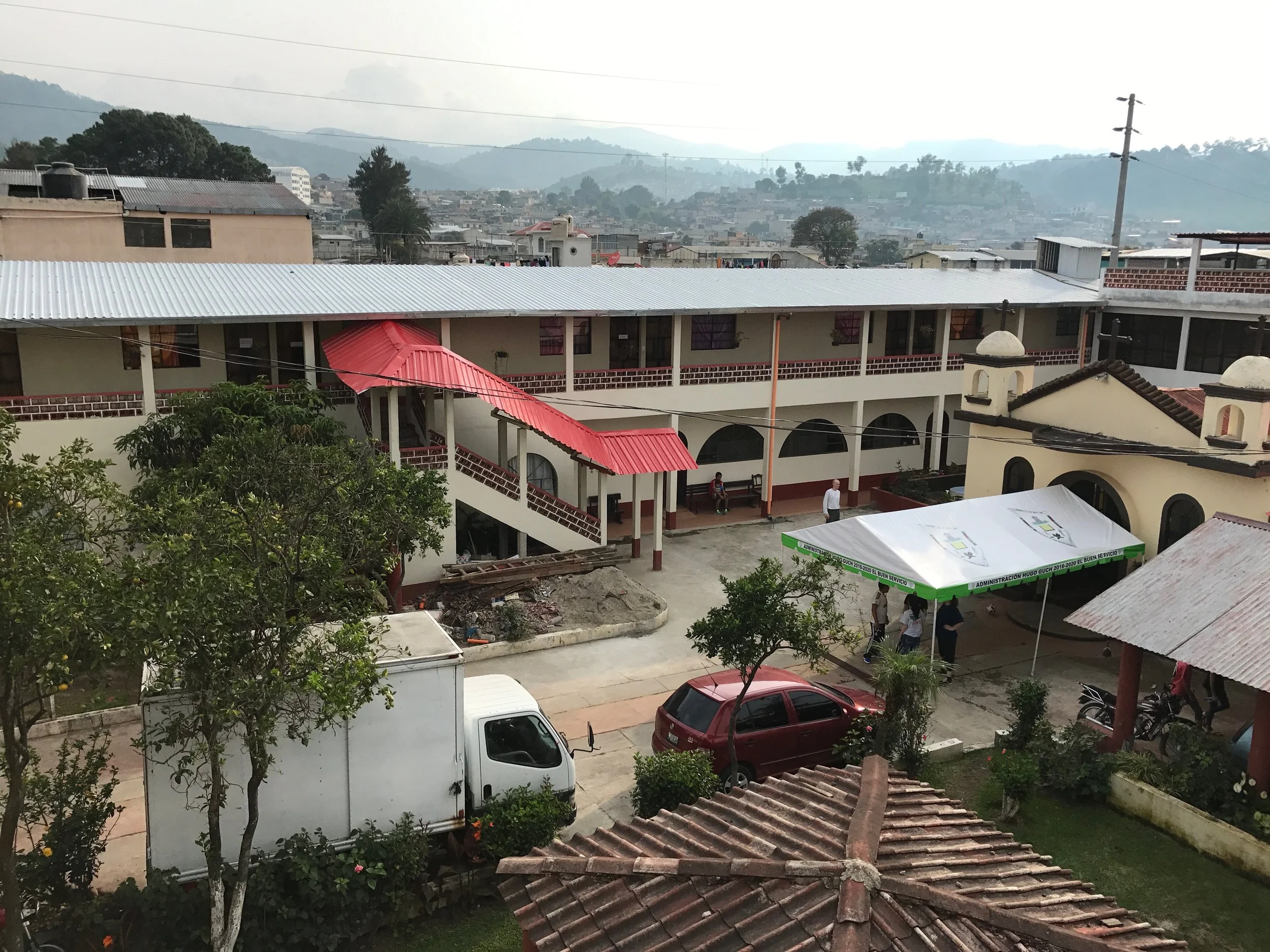 A view of a school campus with two-story buildings, parked cars, trees, and a parking lot. There are people near a tent and on the walkway, with mountains in the background.
