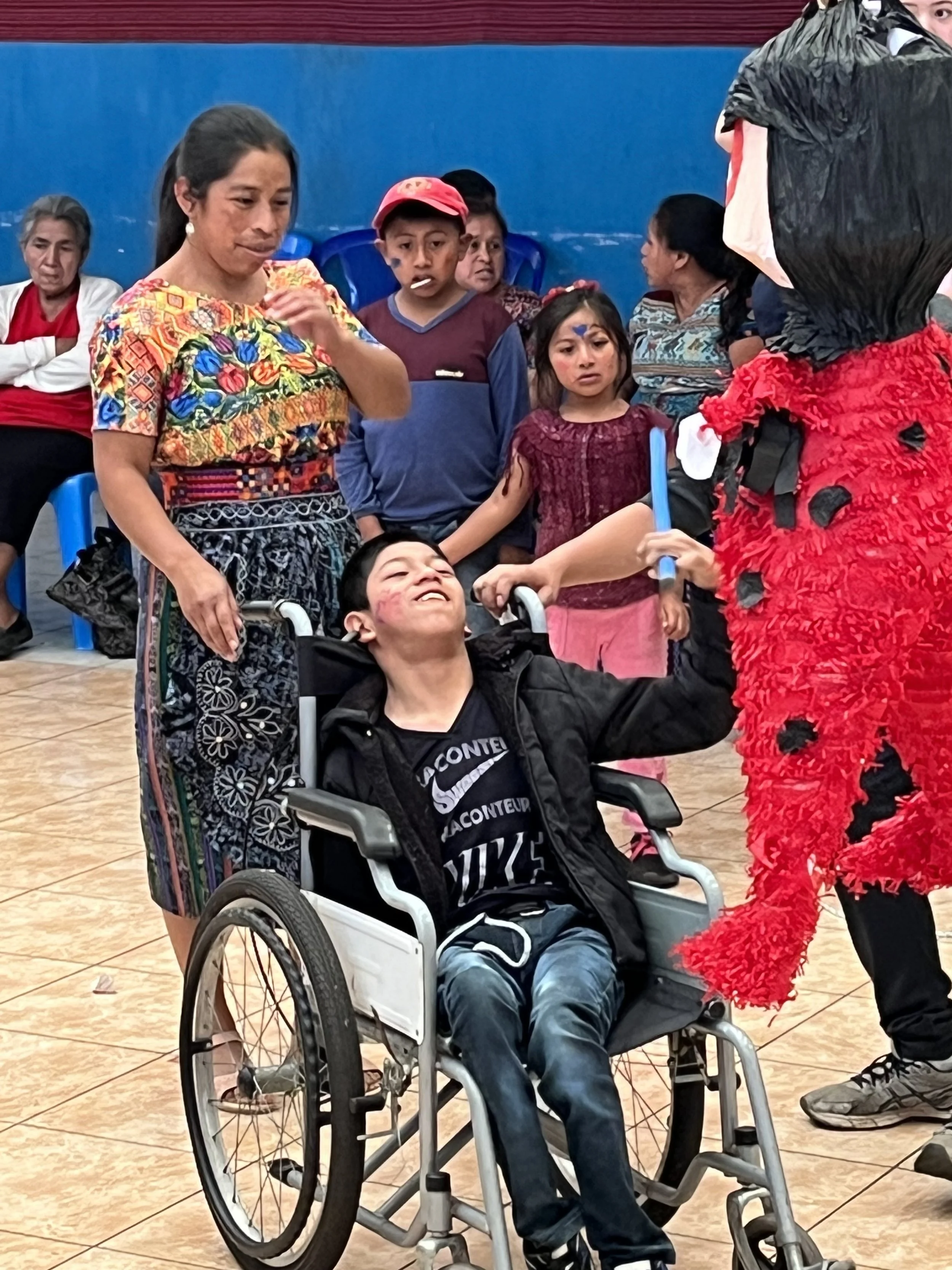 A young man in a wheelchair smiling and raising his arm towards a person dressed in a ladybug costume, while surrounded by several onlookers, including women and children, in a colorful indoor setting.