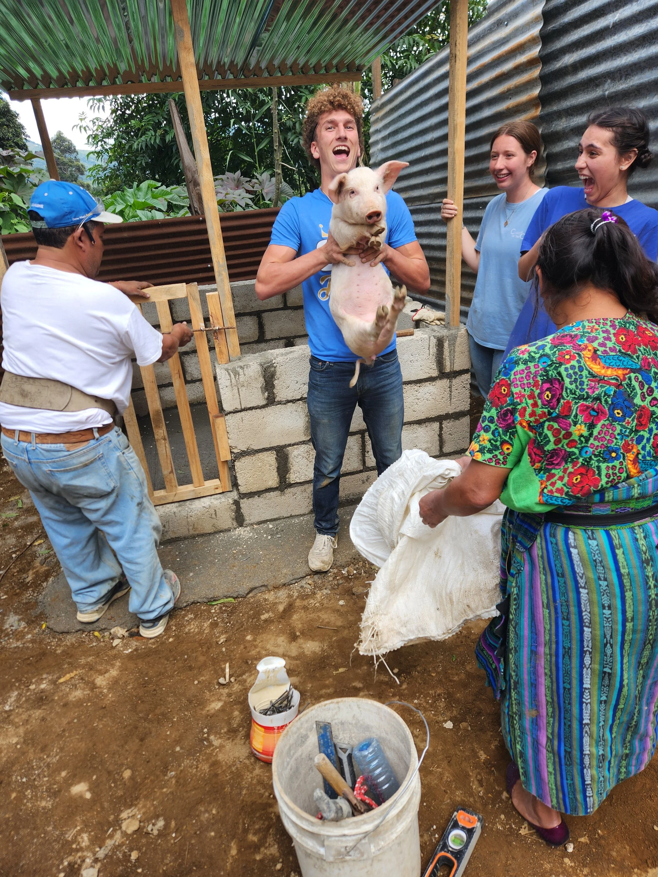 A group of people in a construction or farming setting, with a man holding a pig, surrounded by others smiling and watching. There are tools and construction materials on the ground, and the area looks semi-outdoor with metal and wood roofing.