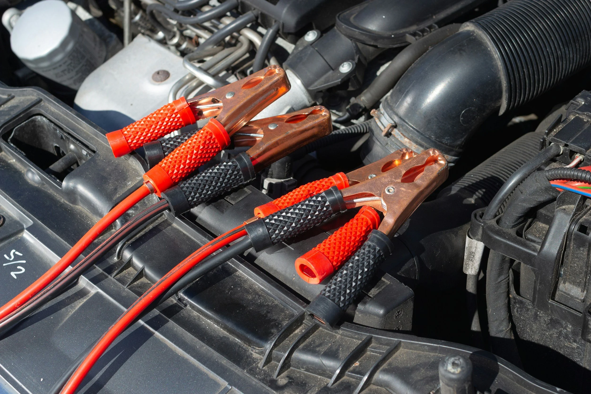 Car battery with four jumper cables attached, situated under the hood of a vehicle