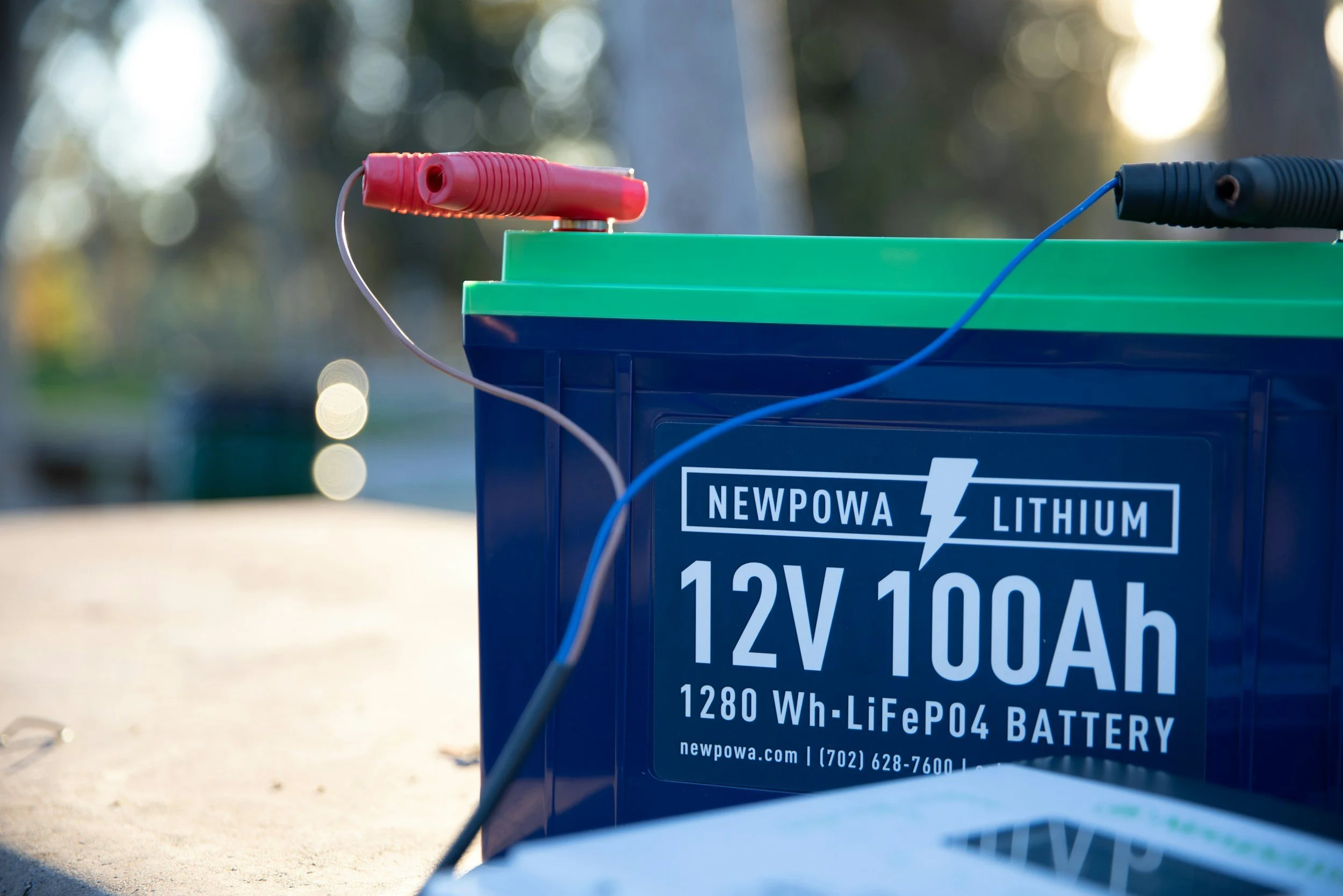 Close-up of a blue lithium-ion battery with green top, labeled 'Newpowa Lithium 12V 100Ah, 1280 Wh liFePO4 Battery' and website and phone number. The battery has red and black cables connected to it, with blurred background and sunlight.