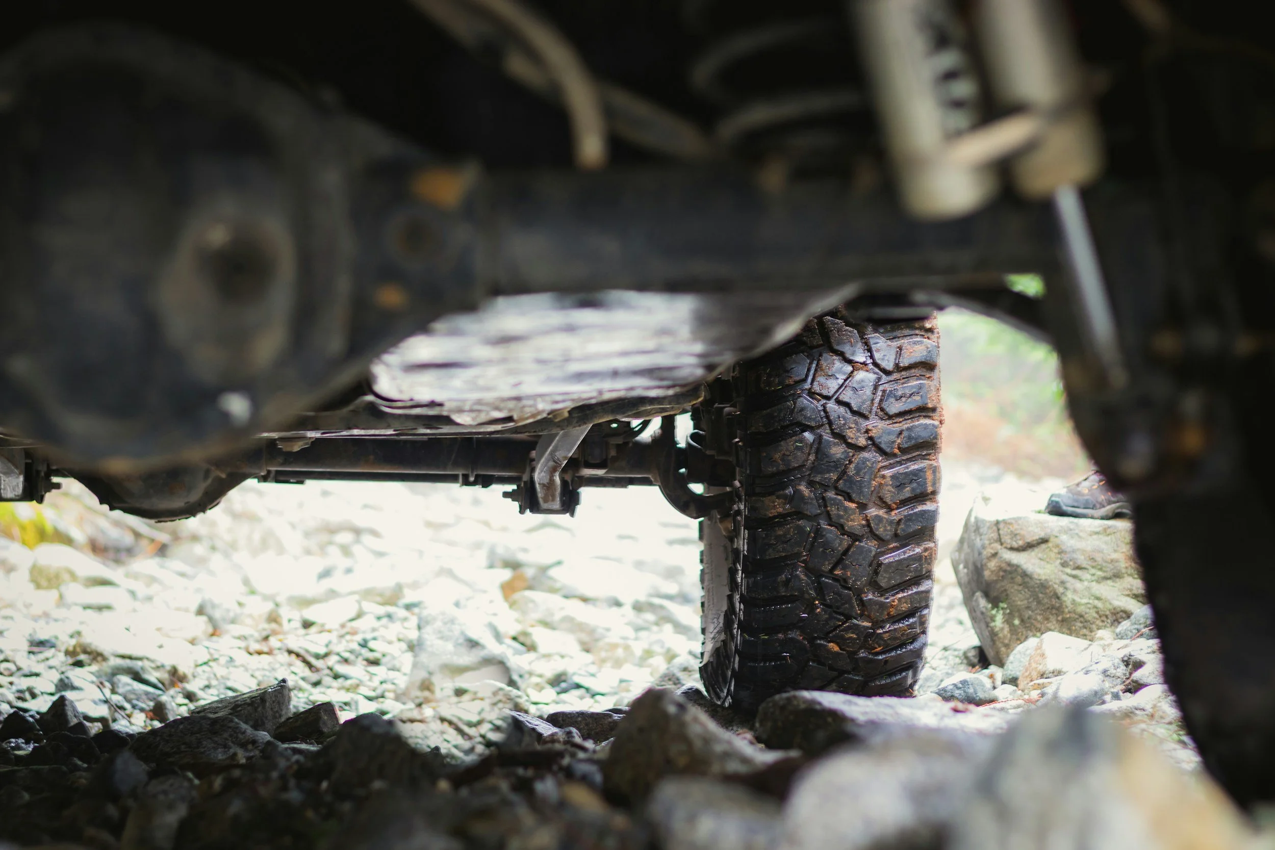 View beneath an off-road vehicle showing a large, rugged tire on rocky terrain.