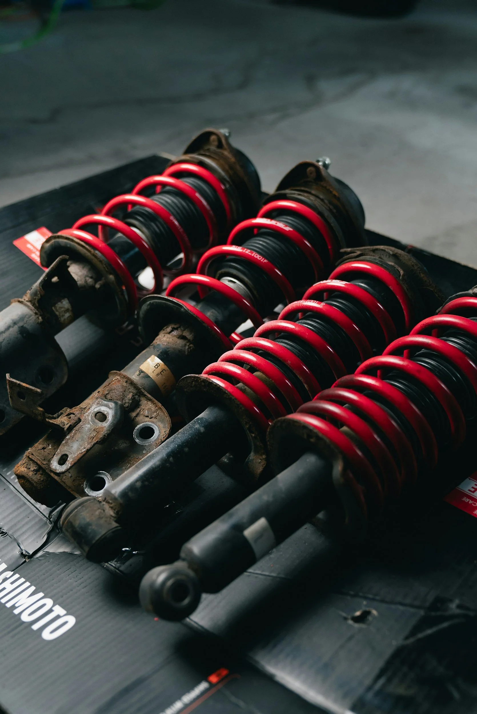 Two sets of used car coilover suspension shocks with red springs laid on a black surface.